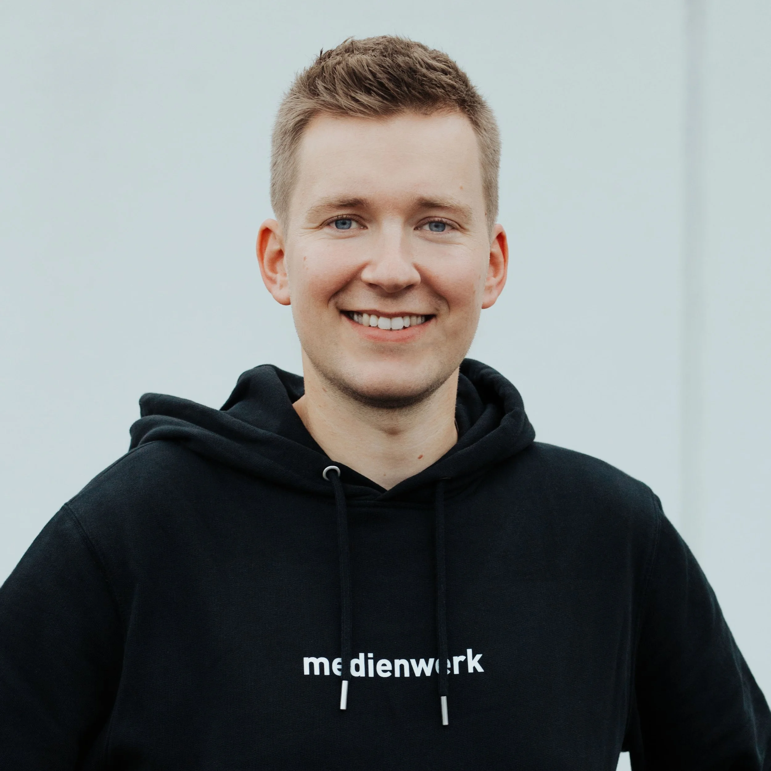Ein junger Mann mit kurzen, hellbraunen Haaren trägt einen schwarzen Hoodie mit weißen Schriftzug 'medienwork' und lächelt vor einem neutralen Hintergrund.