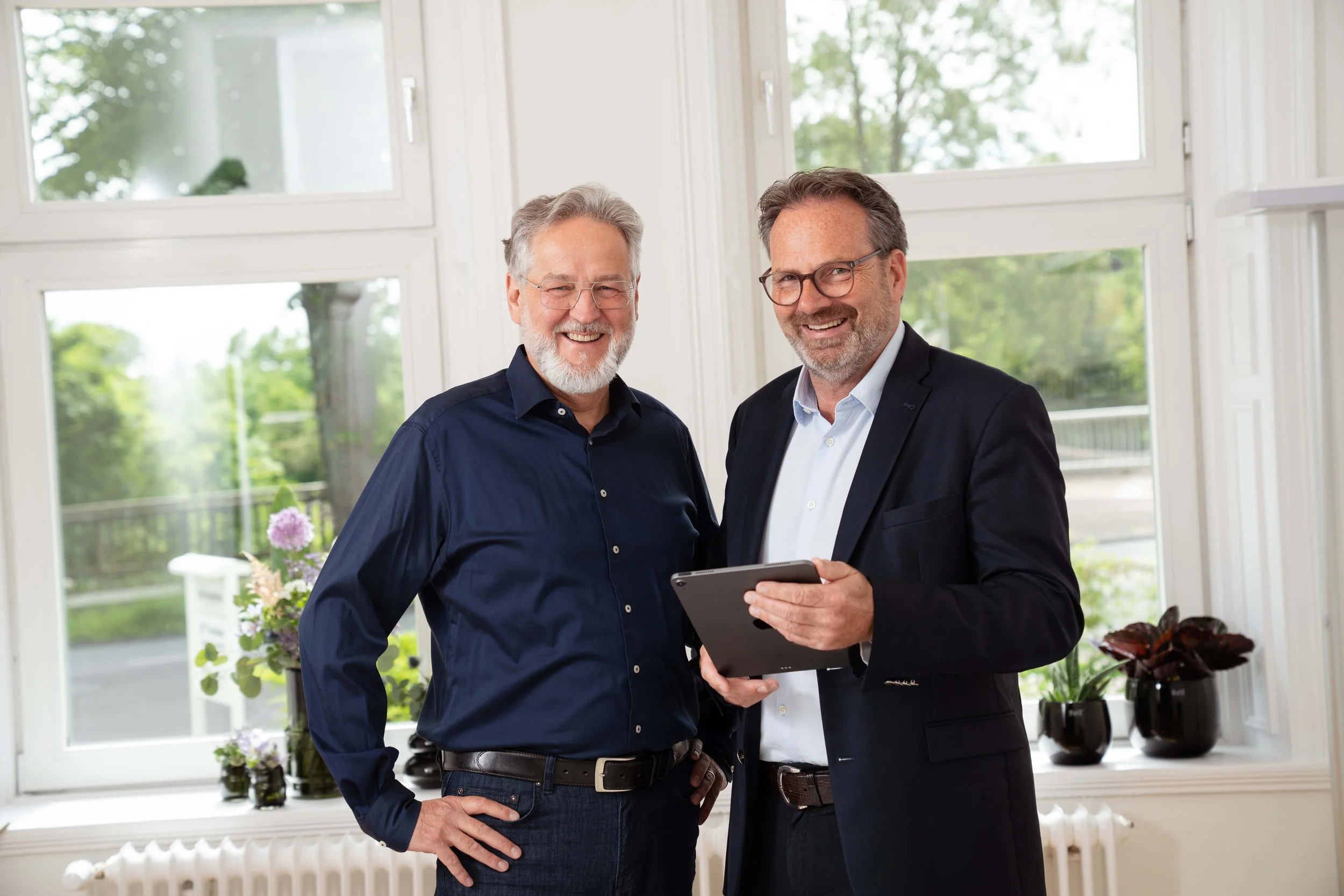 Zwei lachende Männer in formeller Kleidung stehen vor großen Fenstern, einer hält ein Tablet, im Hintergrund sind Pflanzen auf der Fensterbank.