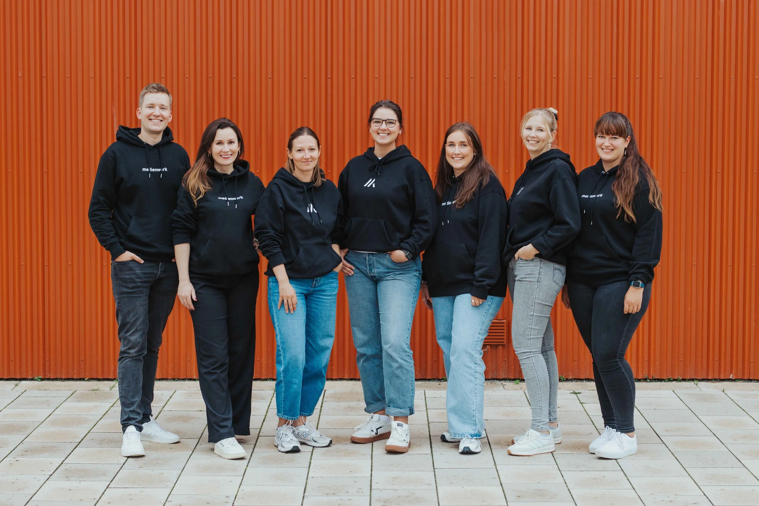 Sieben Personen stehen nebeneinander vor einer orangefarbenen Wand, alle tragen schwarze Hoodies und jeans, lachen und schauen in die Kamera.