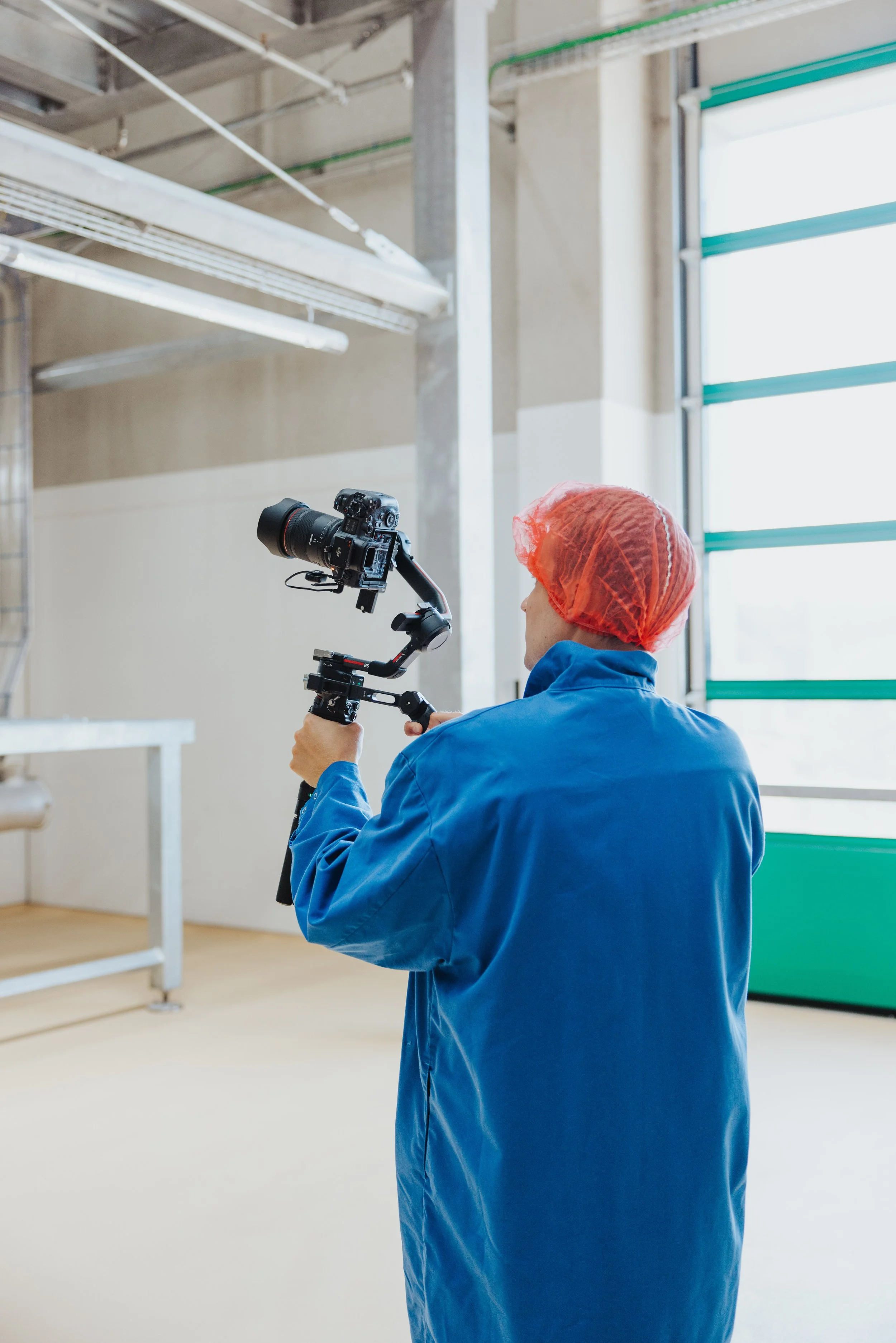 Person in blauer Jacke und rotem Haarnetz filmt mit einer Kamera in einem Industriebetrieb.