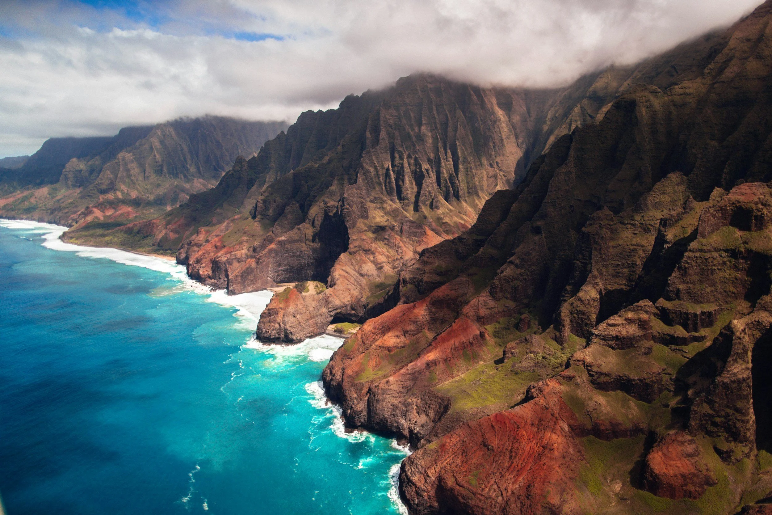 Aerial view of rugged brown and green cliffs along the coast with turquoise blue ocean waves crashing at the base, partly covered by clouds.