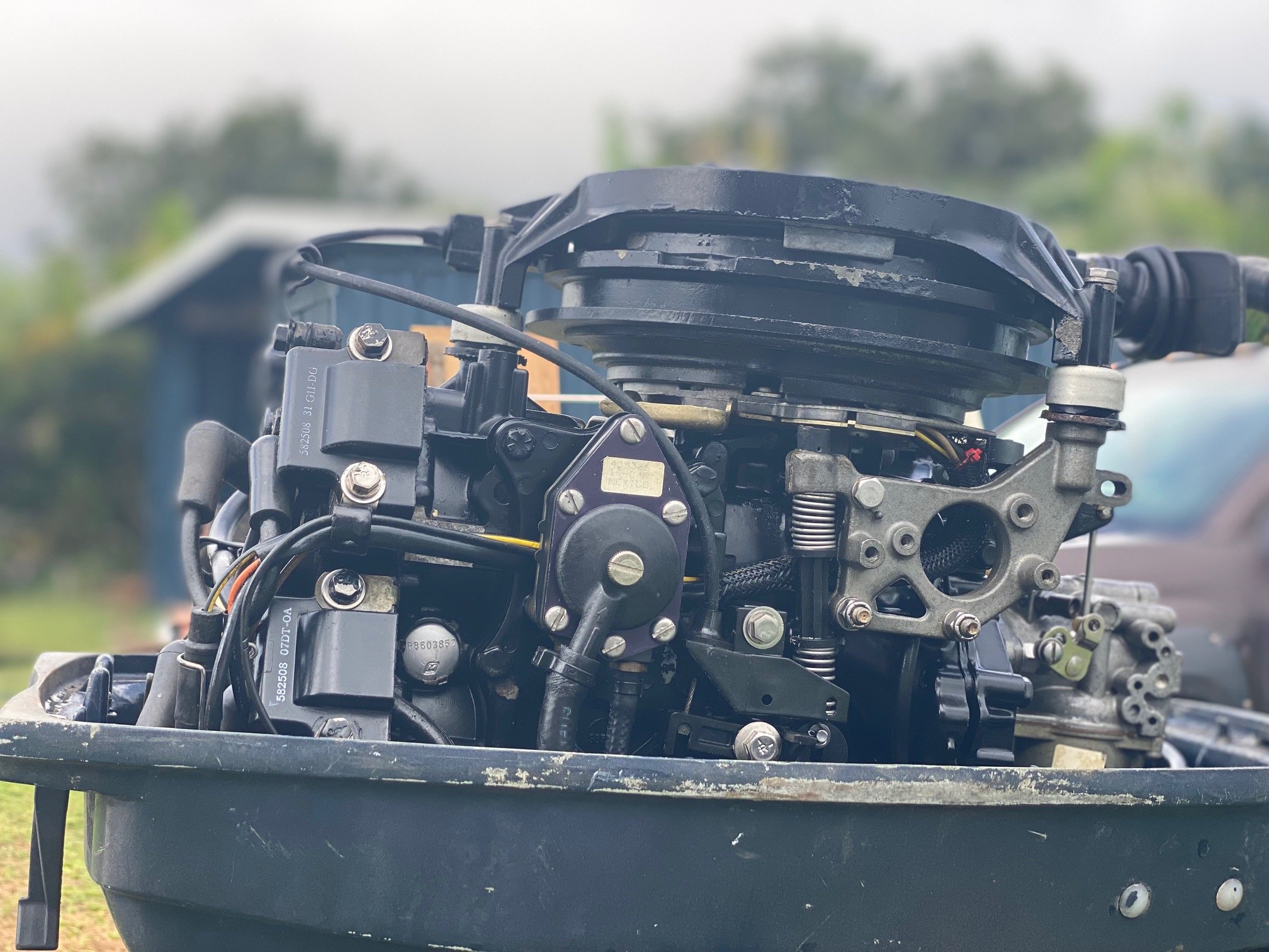 Close-up of an outboard boat motor with visible mechanical components, mounted on a black basin outdoors, blurred background with trees and a vehicle.