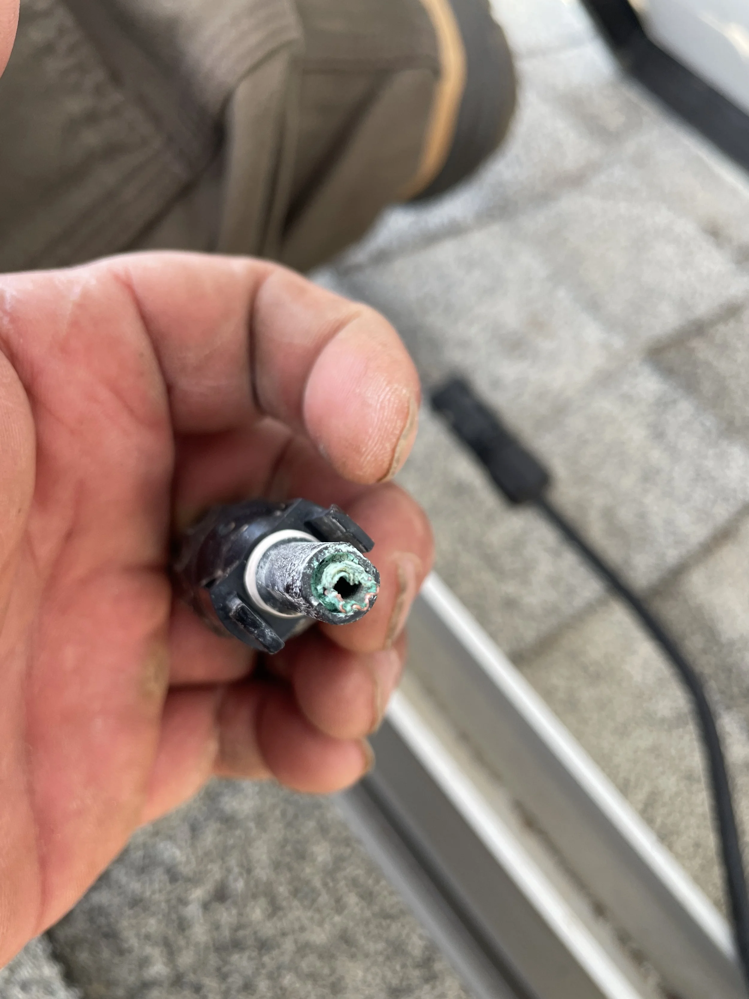 Close-up of a person's hand holding a damaged or corroded electrical connector or sensor, with a blurred outdoor background.