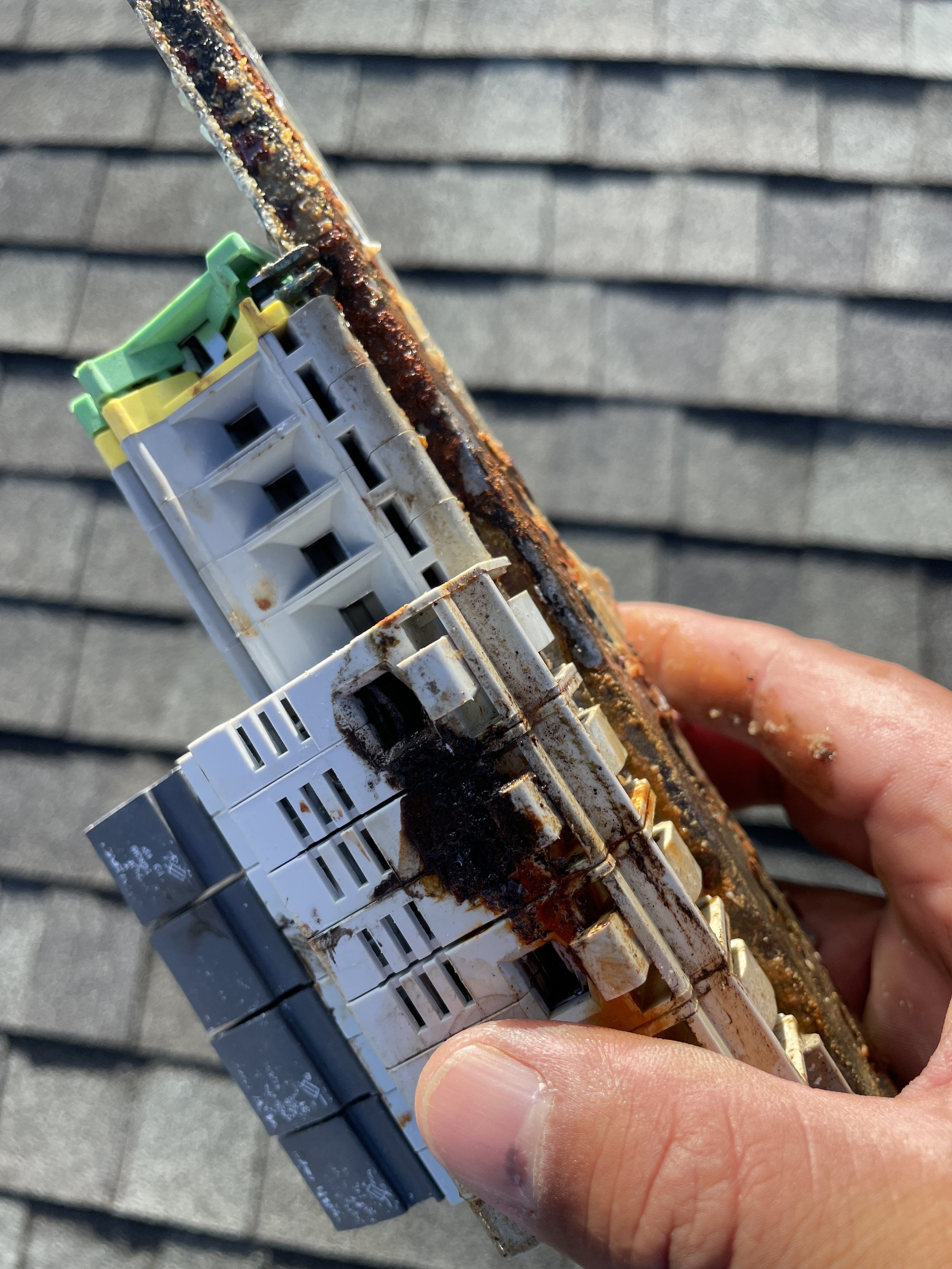 A person's hand holding a rusty electrical component with burnt marks and corrosion, against a background of gray roof shingles.