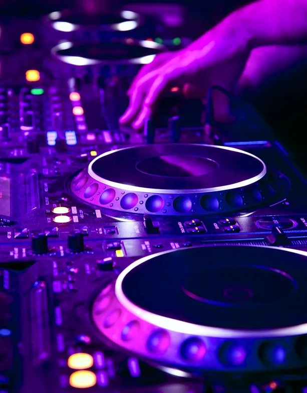 Nahaufnahme eines DJ-Pults mit leuchtenden Lichteffekten und Hand, die einen Knopf bedient, in einer dunklen, neonbeleuchteten Umgebung.