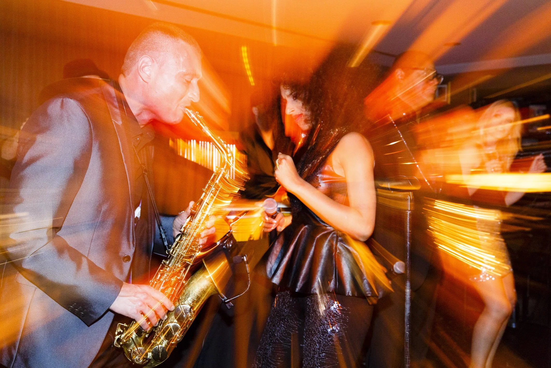 Menschen bei einer Party, einer spielt Saxophon, eine singt, alle in festlicher Stimmung, bunte Lichter im Hintergrund, Bewegungsunschärfe durch Tanz.