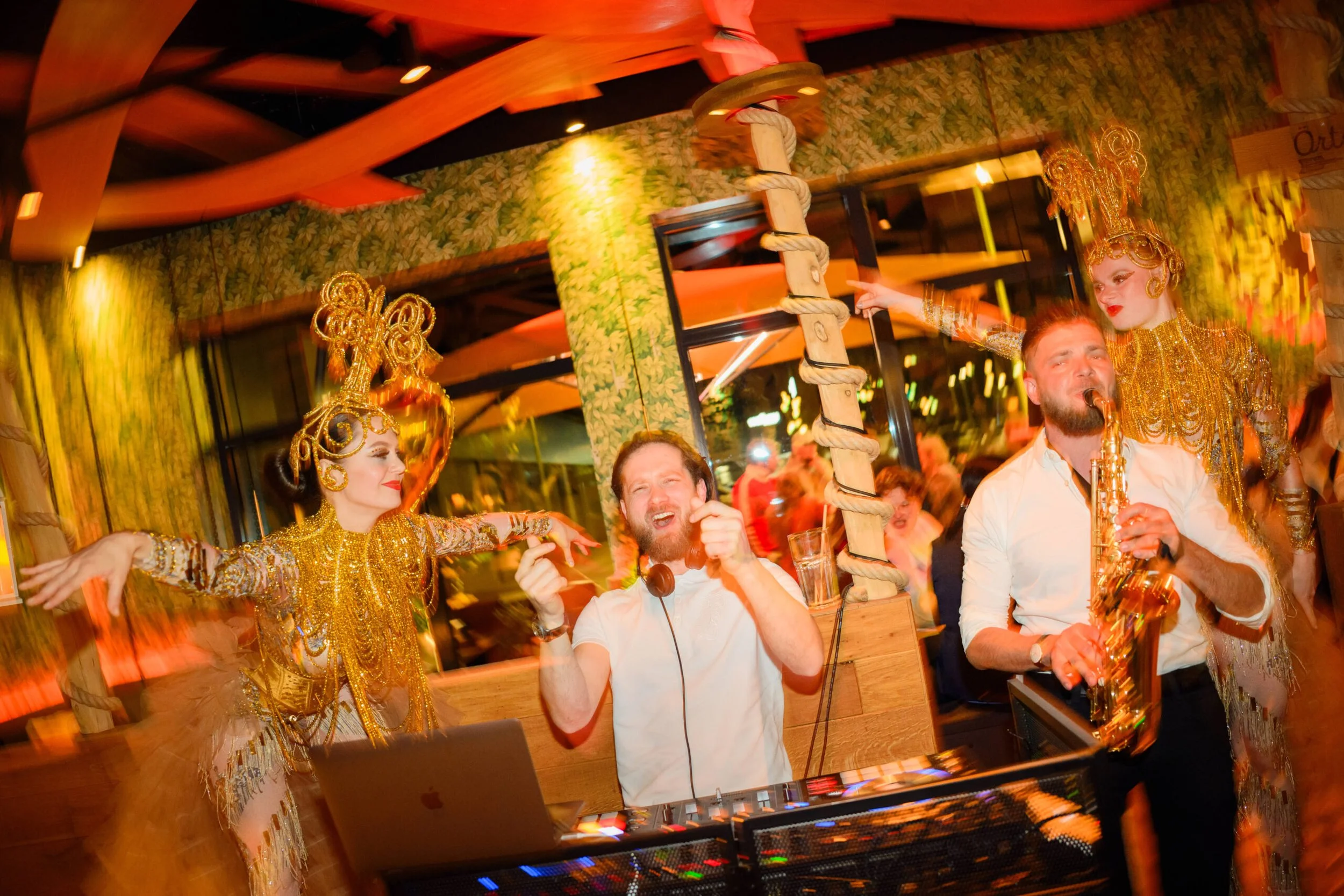 Eine Party mit Menschen, die sich mit goldenen Kostümen und Accessoires verkleidet haben, im Hintergrund bunte Wanddekoration und eine Diskokugel.