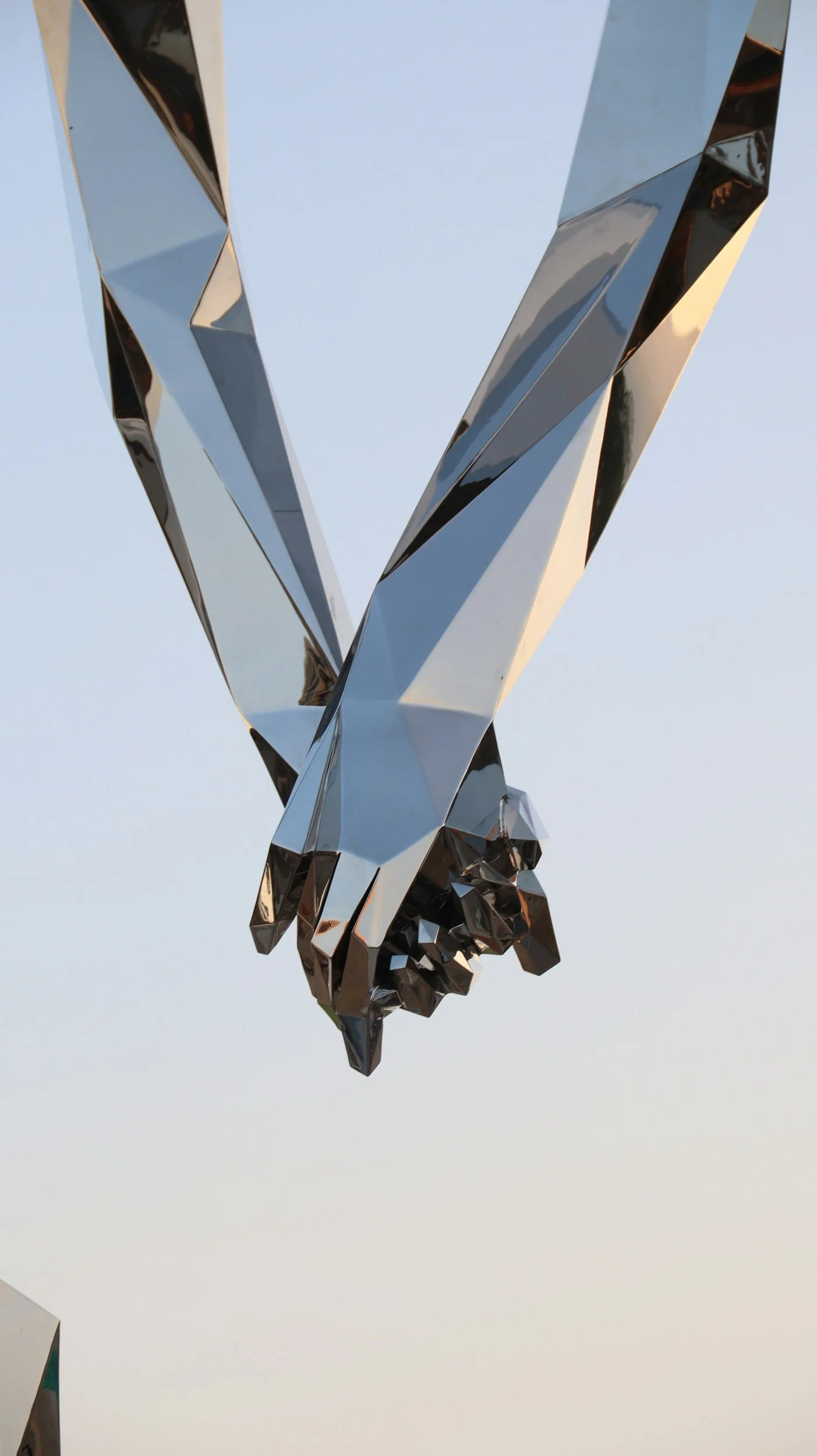 Close-up of a reflective metallic sculpture of two hands holding each other against a light sky background.