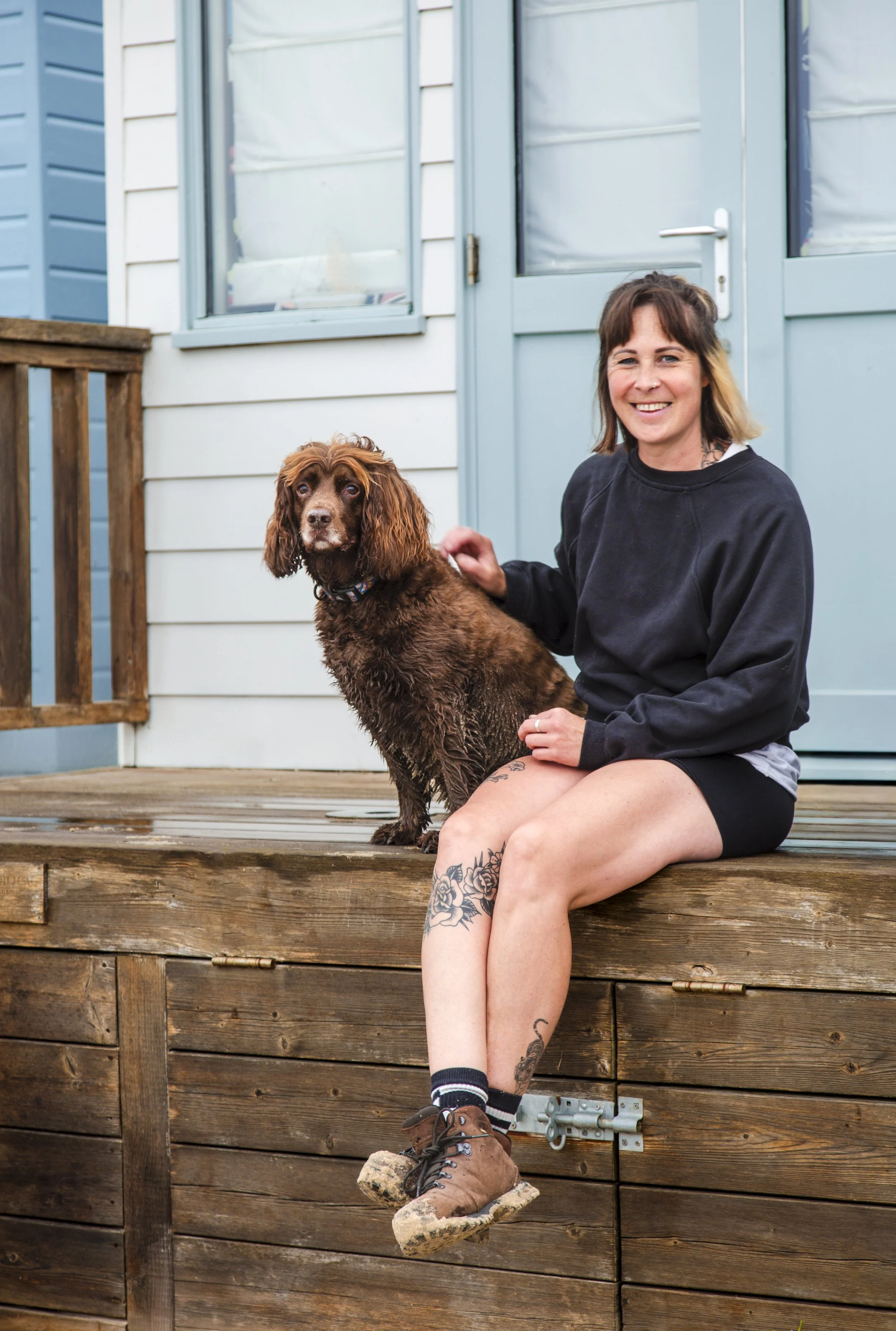 A young woman with tattoos on her leg and wearing hiking boots is sitting on a wooden deck with her hand on a brown spaniel dog. She is smiling, and they are in front of a blue house with white siding and a screen door.