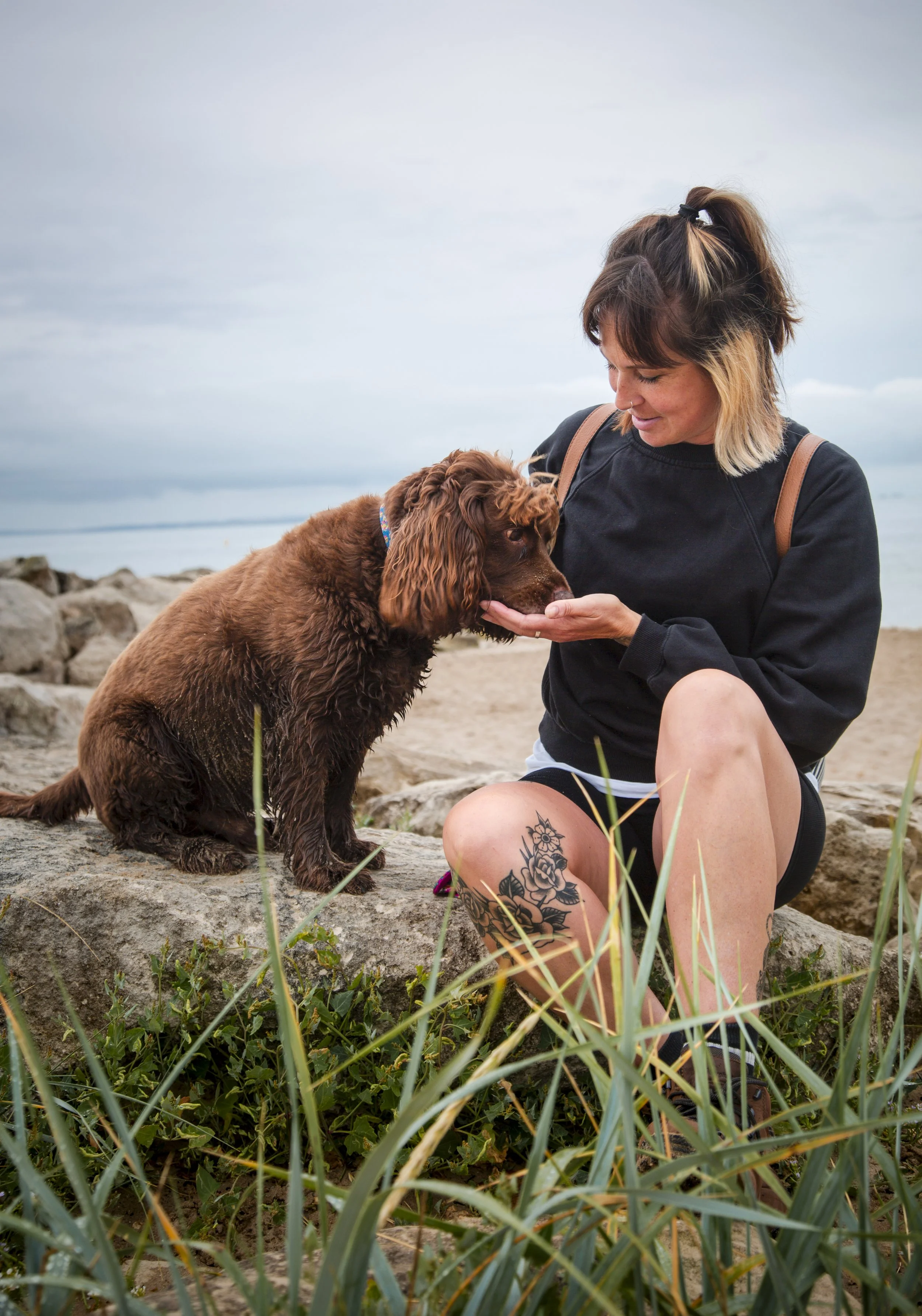 A woman with short hair and a tattoo on her leg, smiling while holding her dog's face at a beach with rocks, grass, and cloudy sky in the background.