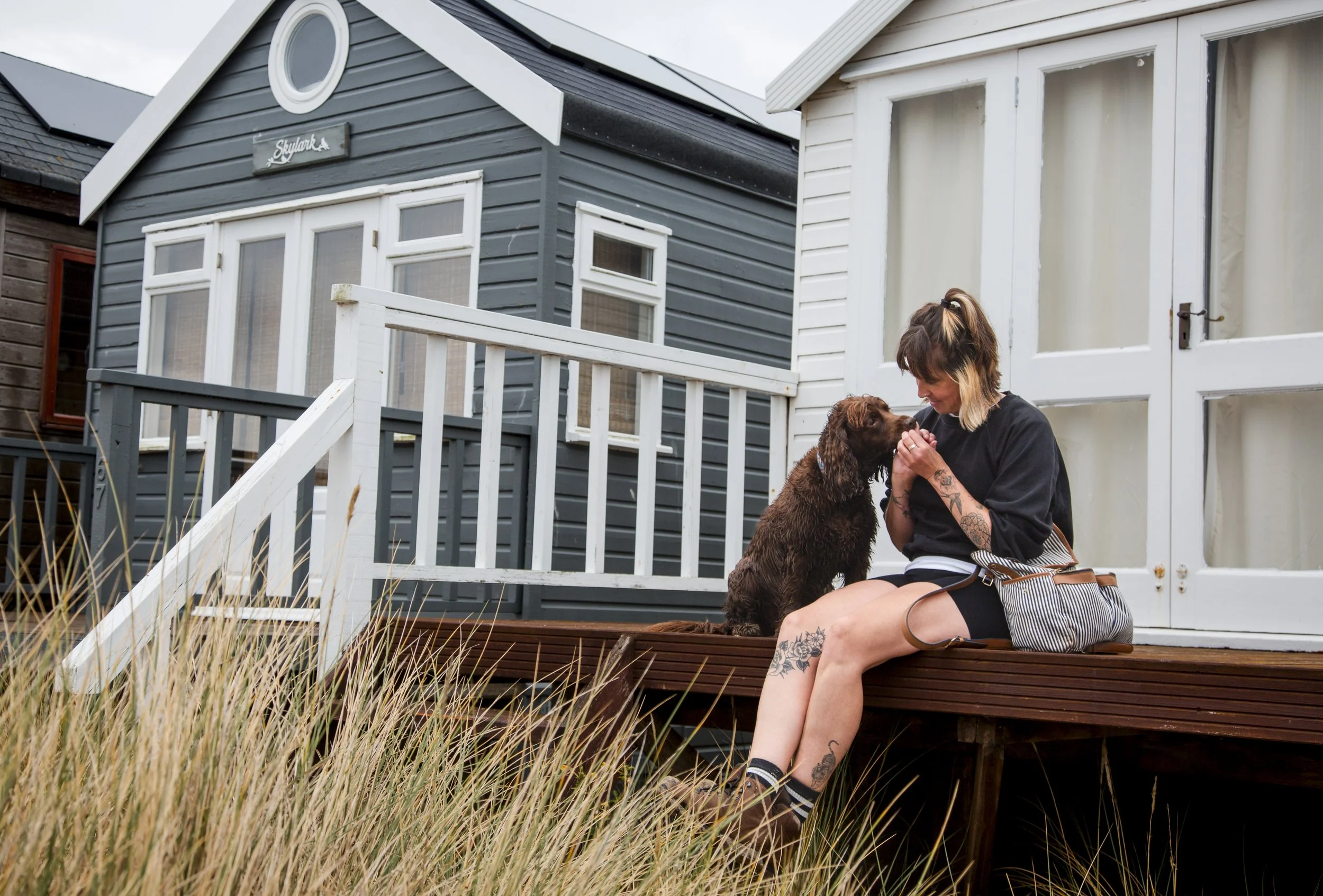 A woman with tattoos and short hair sitting on a porch, playing with a brown puppy. The porch is attached to a neighborhood house with gray and white siding and a white door.