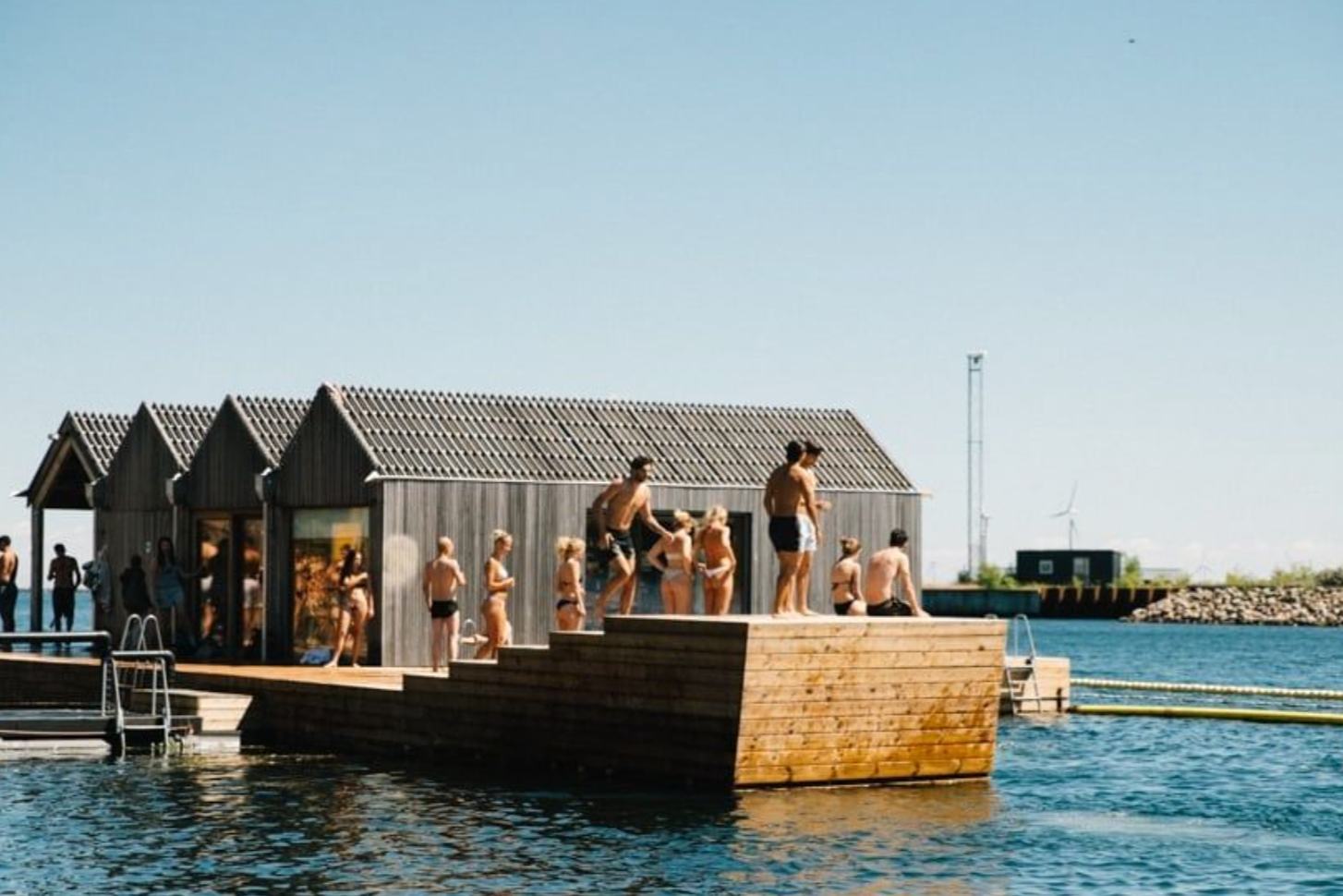 People in swimsuits preparing to jump into the water from a wooden dock near a small wooden building on a clear, sunny day.