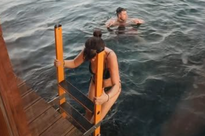 A woman in a swimsuit climbing out of a body of water using a pool ladder, while a man in a swimsuit is in the water nearby.