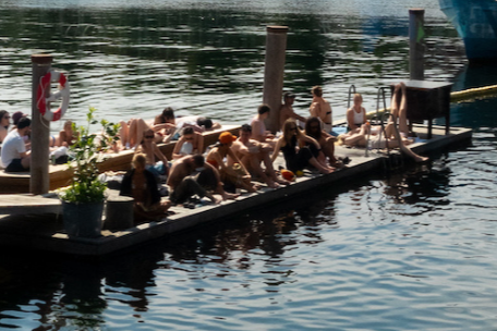 People sunbathing and sitting on a dock by the water, some with umbrellas and a piano at the end of the dock.