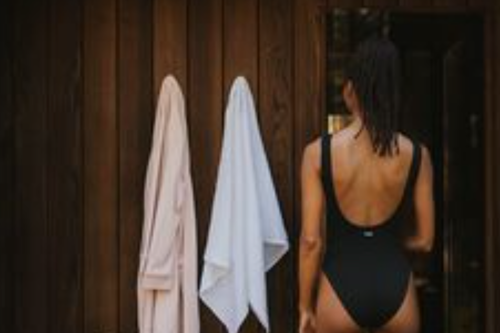 Woman with shoulder-length hair in a black swimsuit looking at a mirror, with two towels hanging on a wooden wall