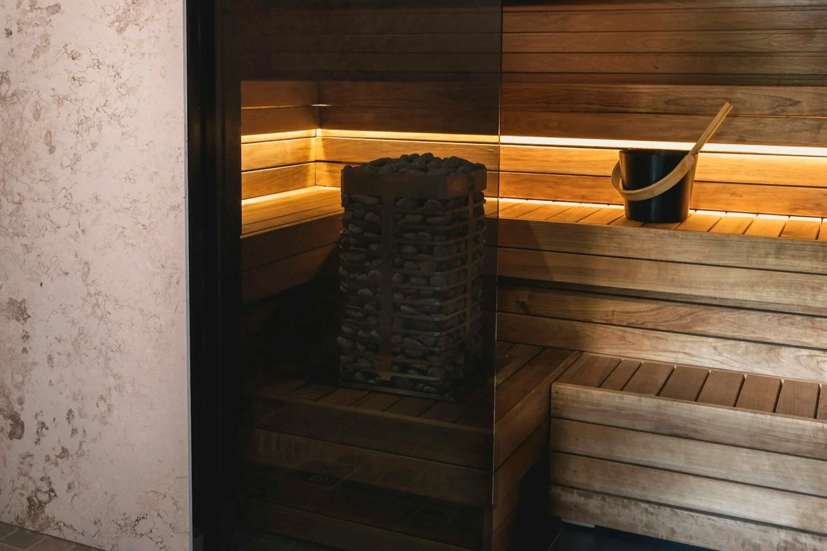 Interior of a traditional sauna with wooden benches, a bucket with a ladle, and a heating element filled with stones.