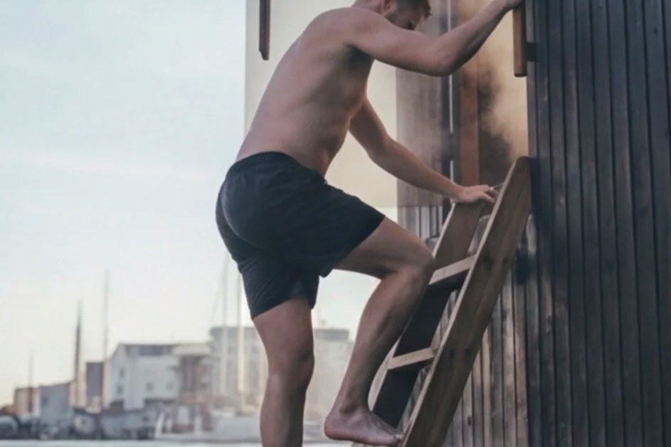 A shirtless man with dark hair wearing black shorts climbing a wooden ladder against a dark wooden wall outside.