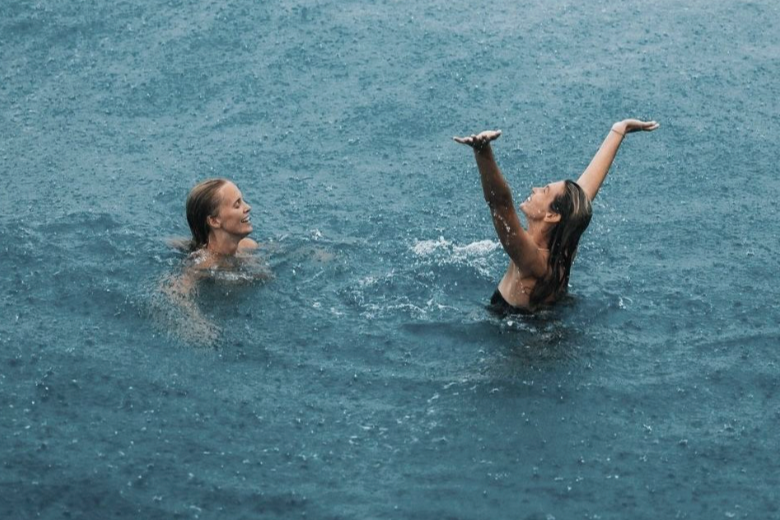 Two women swimming and playing in a large body of water, with one woman smiling and the other raising her arms in a joyful gesture.