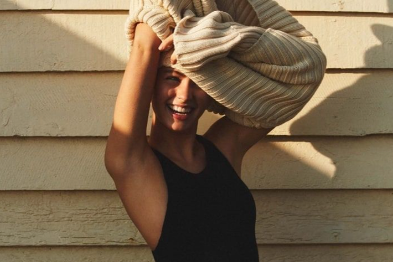 Smiling woman with short hair wearing a black tank top, balancing a large beige striped bag on her head, standing in front of a beige wooden wall.