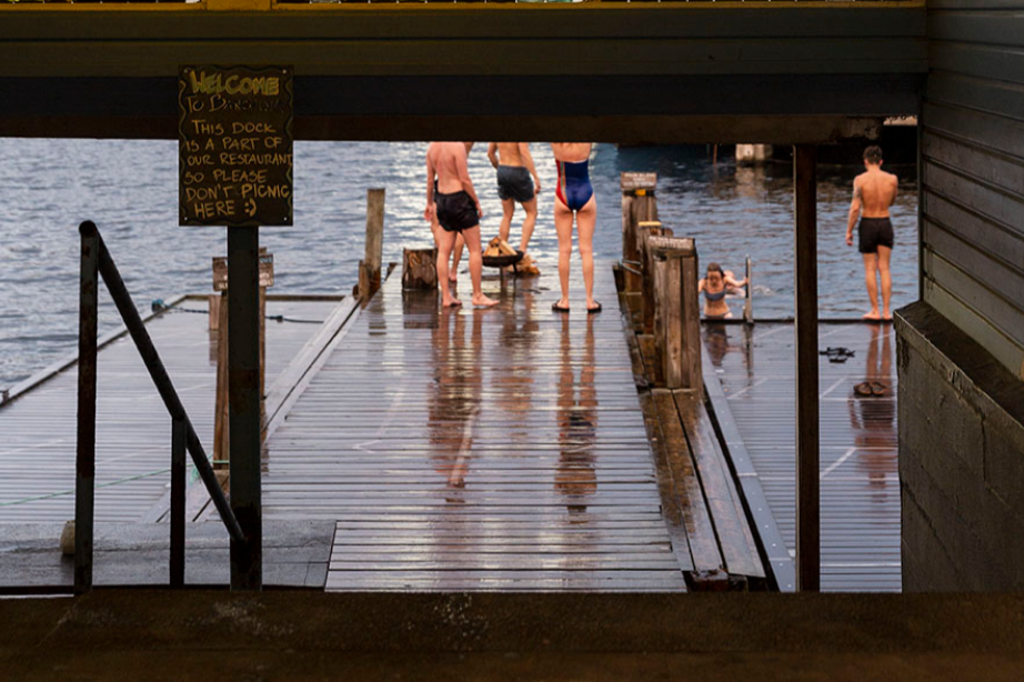 Group of people swimming and standing on a wooden dock by the water, view from under a bridge with a sign that reads welcoming visitors and advising against picnicking.