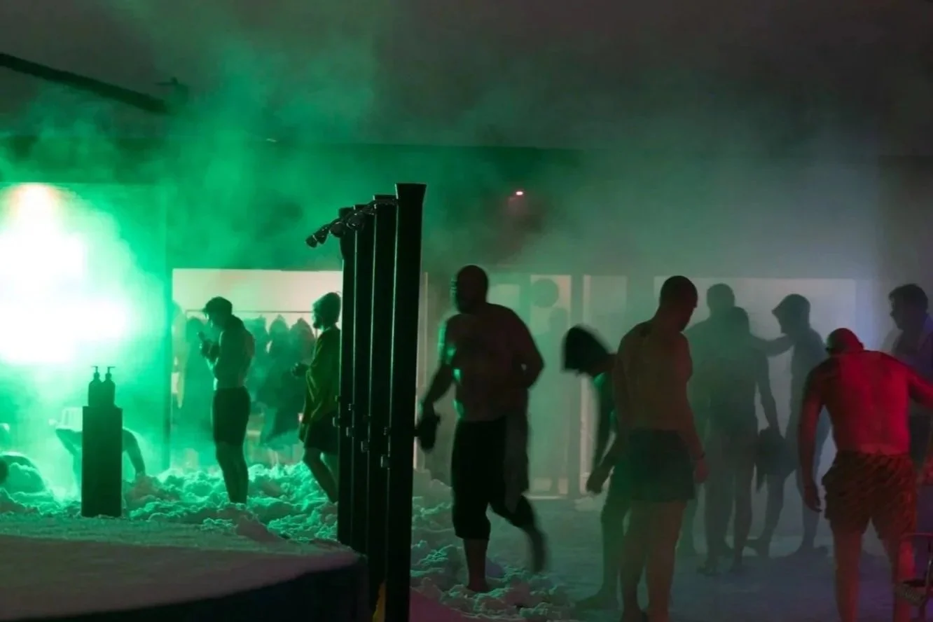 People enjoying themselves at a party with colorful green and red lights, surrounded by smoke or fog, and a foam-covered floor.