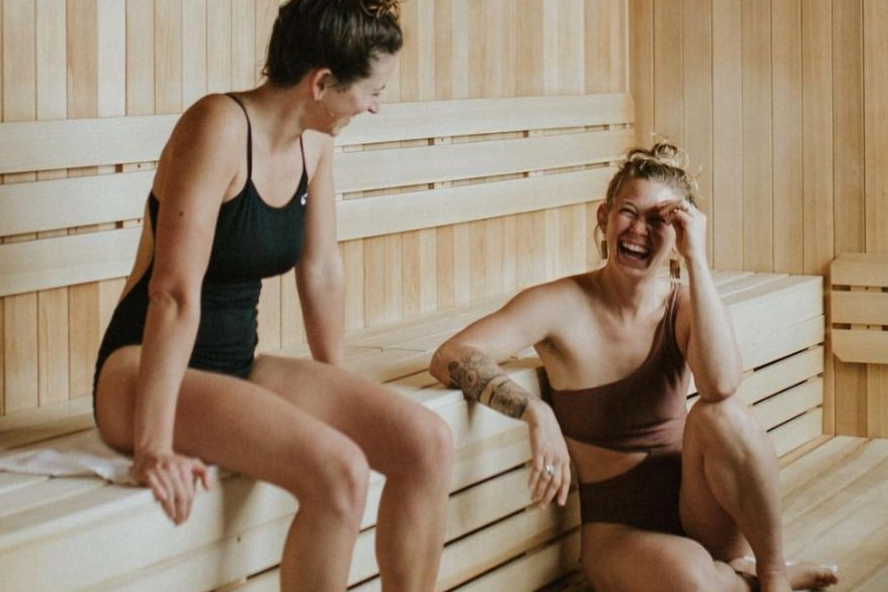 Two women laughing and sitting on a wooden bench in a sauna, wearing swimsuits.