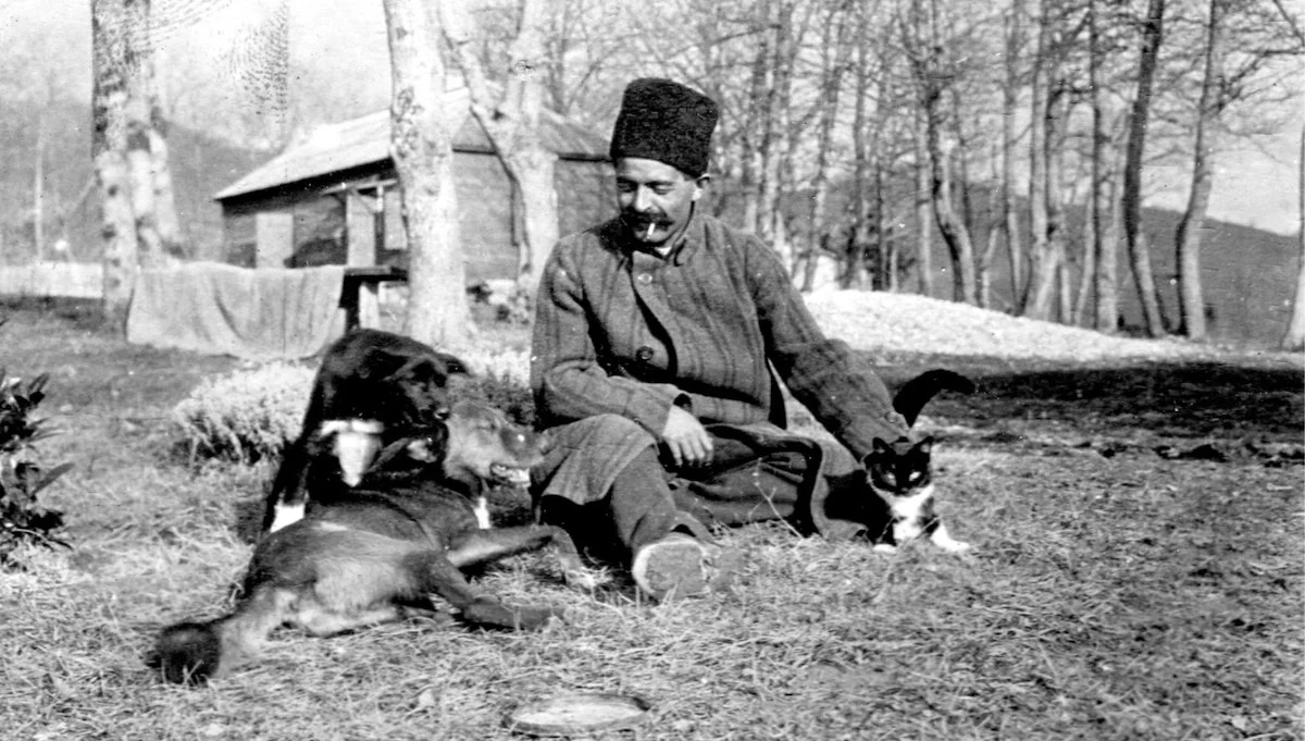 G. I. Gurdjieff, sitting outdoors on the ground with a dog and a cat, in a rural area with trees and a wooden building in the background.