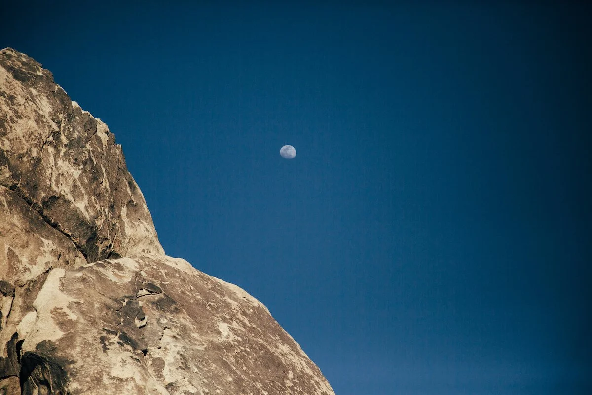 Nighttime scene showing a large rocky mountain with the moon visible in a clear blue sky.