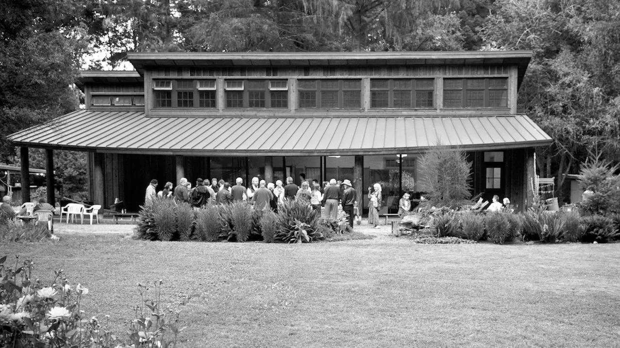 People gathered outside a large house or lodge with a metal roof and porch, surrounded by bushes and trees.