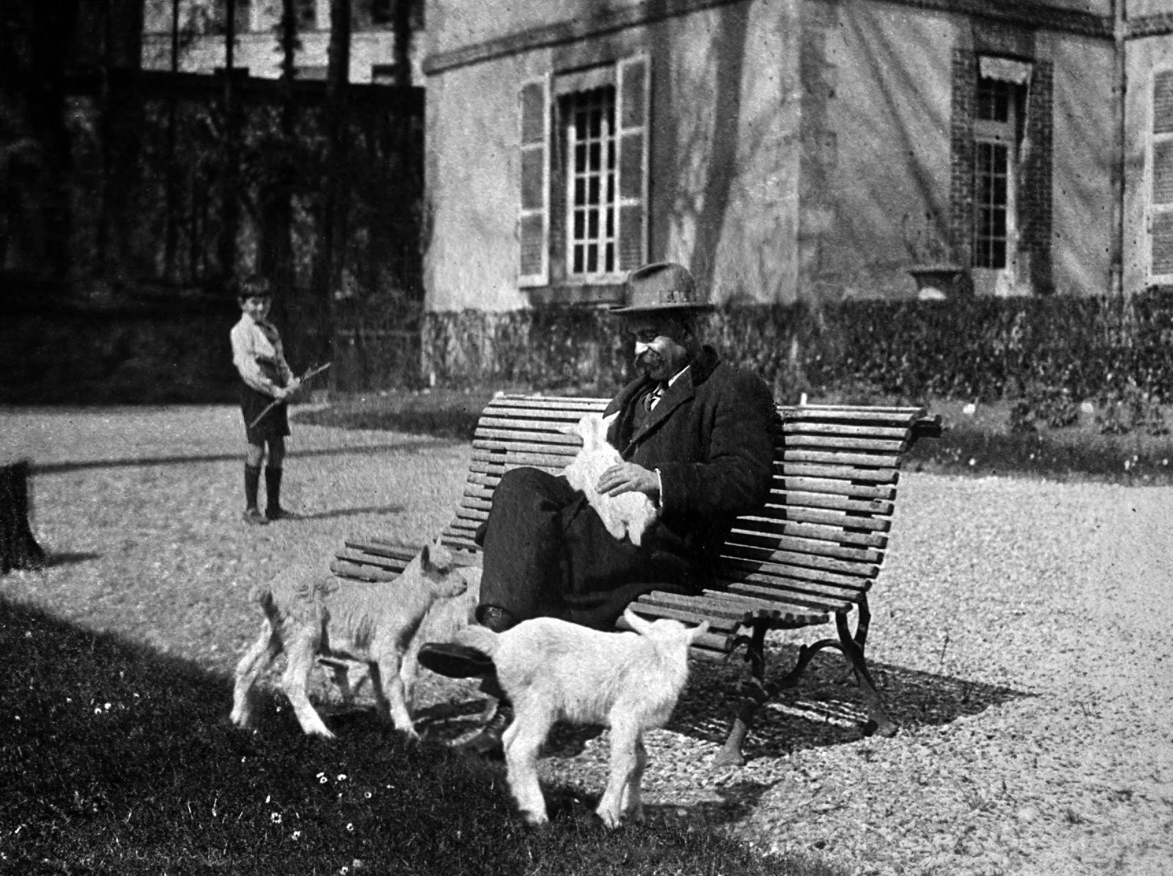 An elderly man with a beard, wearing a suit and hat, sitting on a park bench, petting a dog. Two other dogs are nearby. A young boy holding a stick stands in the background near trees and a house.