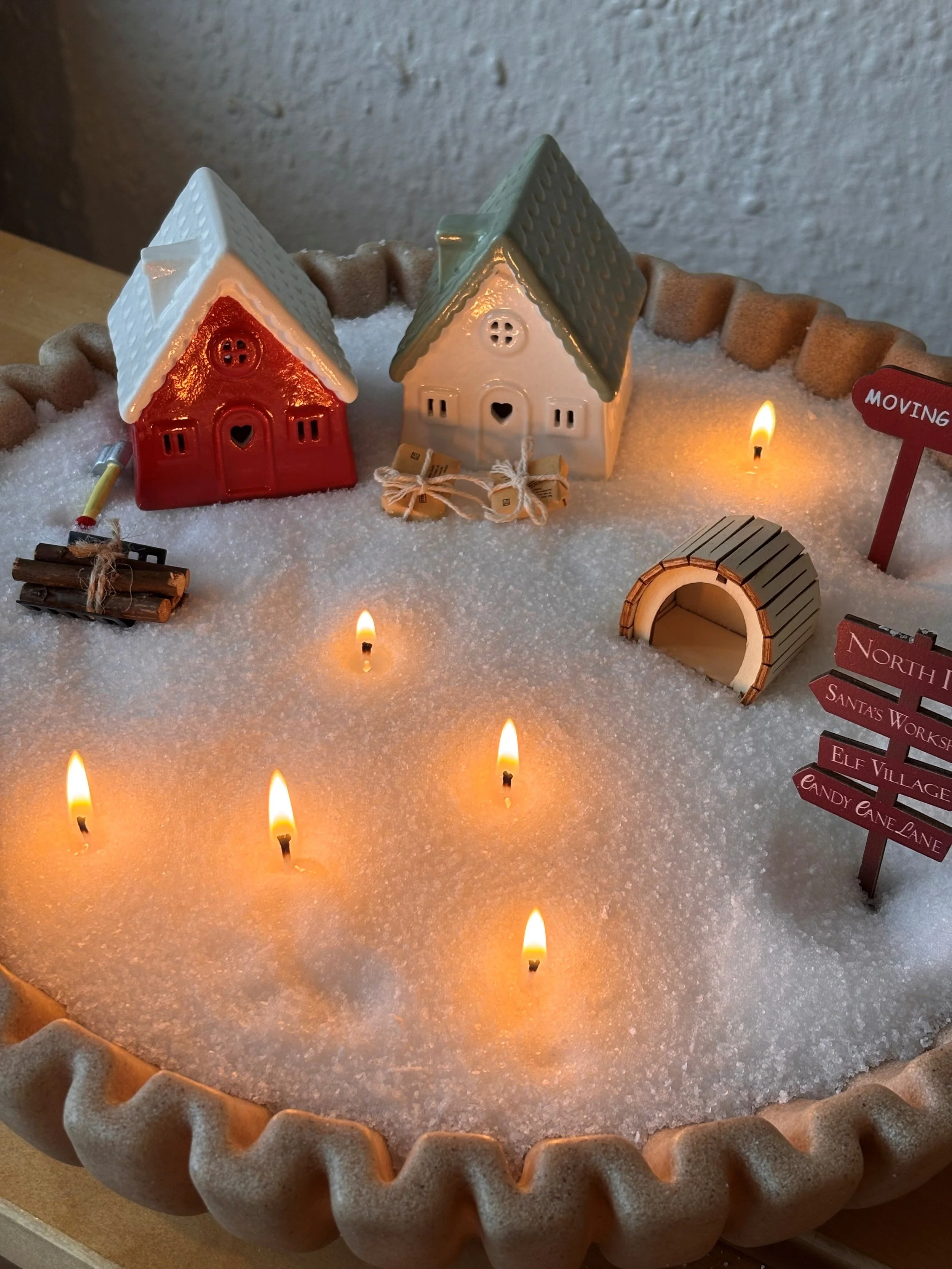 Miniature winter scene with toy houses, small signs, candles, and candy cane lane, all set on artificial snow in a large tart pan.