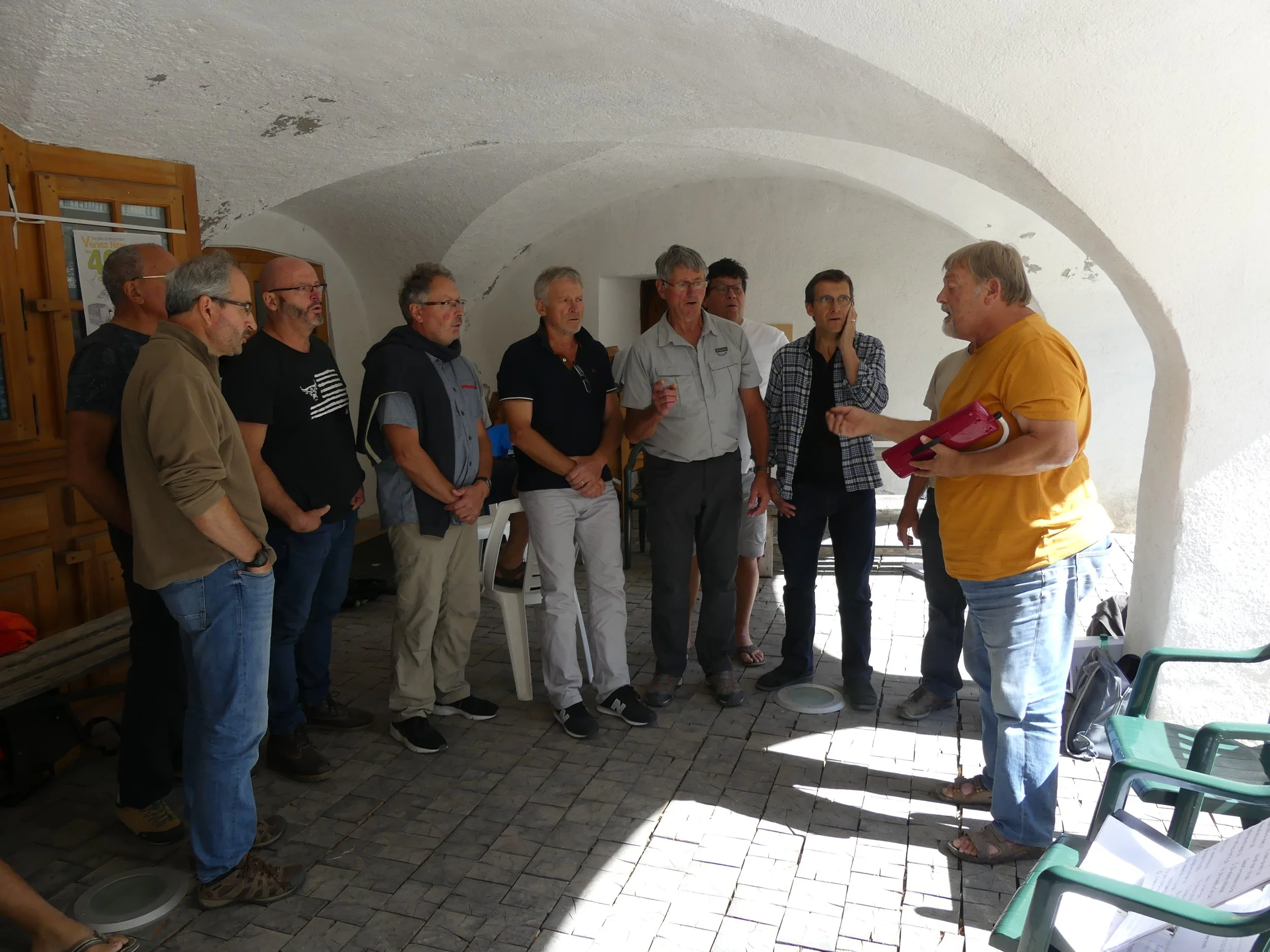 Un groupe d'hommes écoute un homme en jaune qui parle, dans une pièce avec plafond voûté en pierre