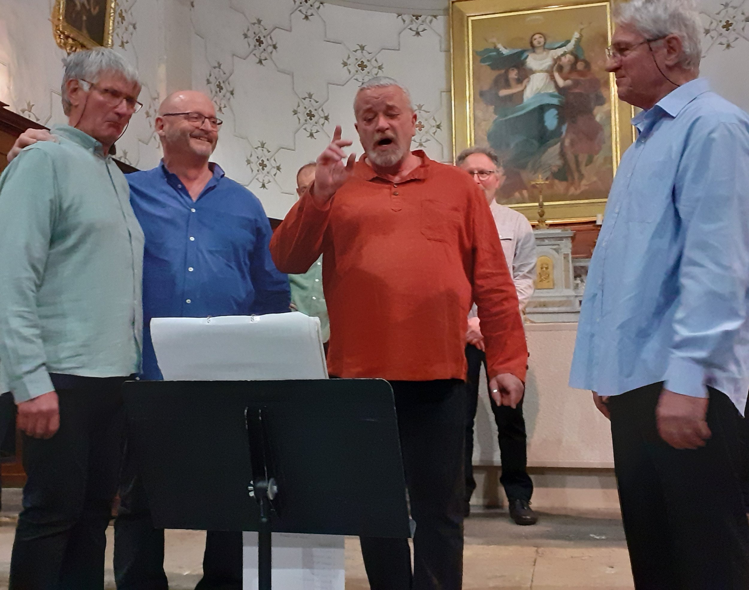 Un groupe de sept hommes, certains avec des lunettes, entourent un homme en rouge qui semble chanter ou parler avec expressivité dans une église ou une salle de restande avec une peinture religieuse en arrière-plan.