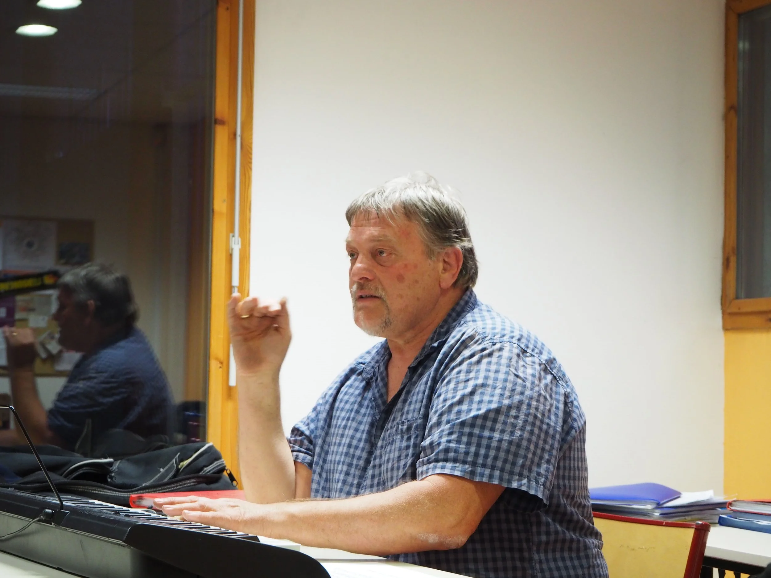 Un homme d'âge moyen joue du piano dans une salle, avec des livres et des dossiers empilés sur une table à côté de lui.