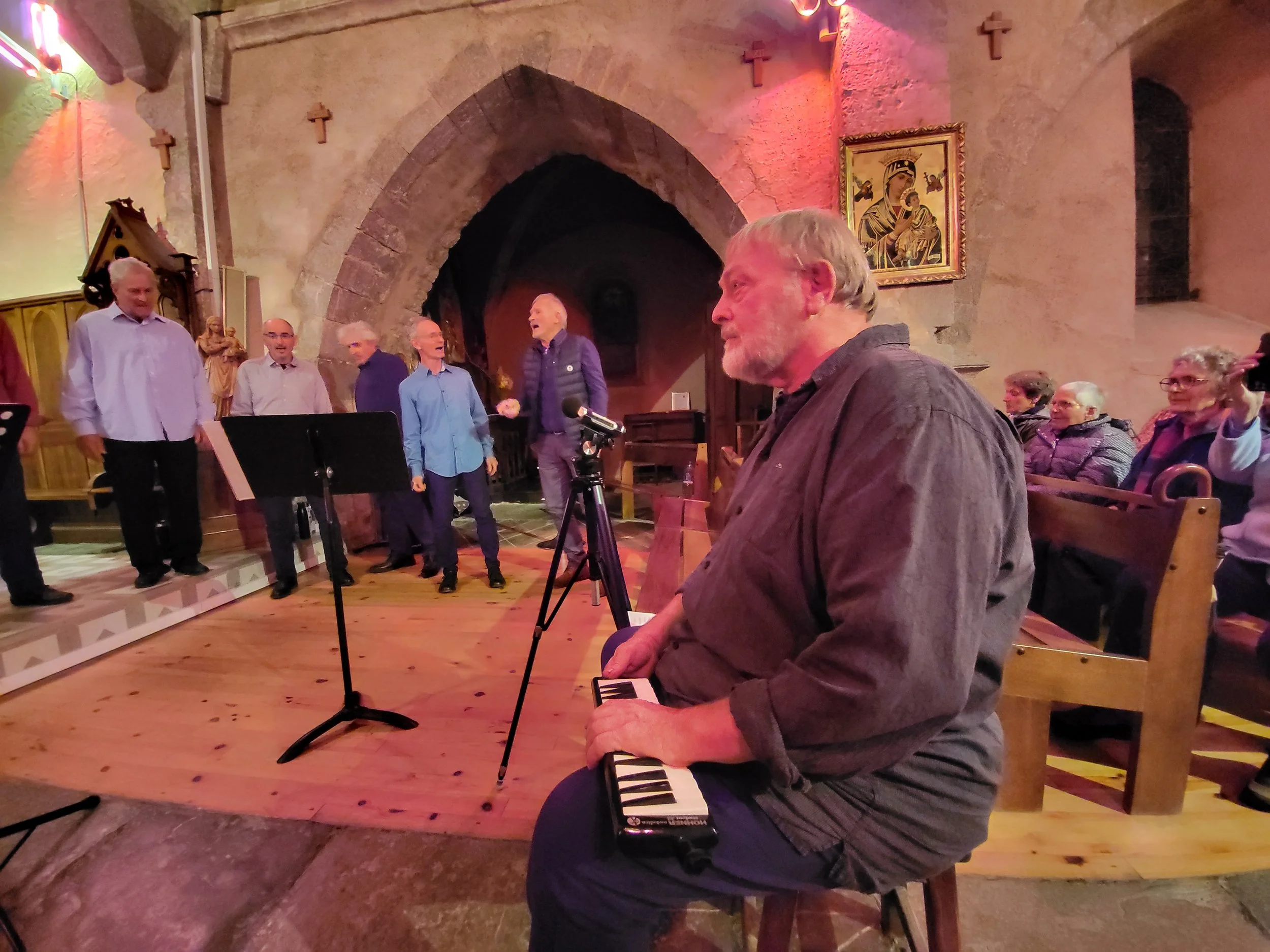 Un homme assis sur une chaise dans une église, jouant du clavier, observant un groupe de personnes chantant ou parlant devant l'autel. À l'arrière, il y a des sièges occupés par d'autres personnes et des décorations religieuses sur les murs.