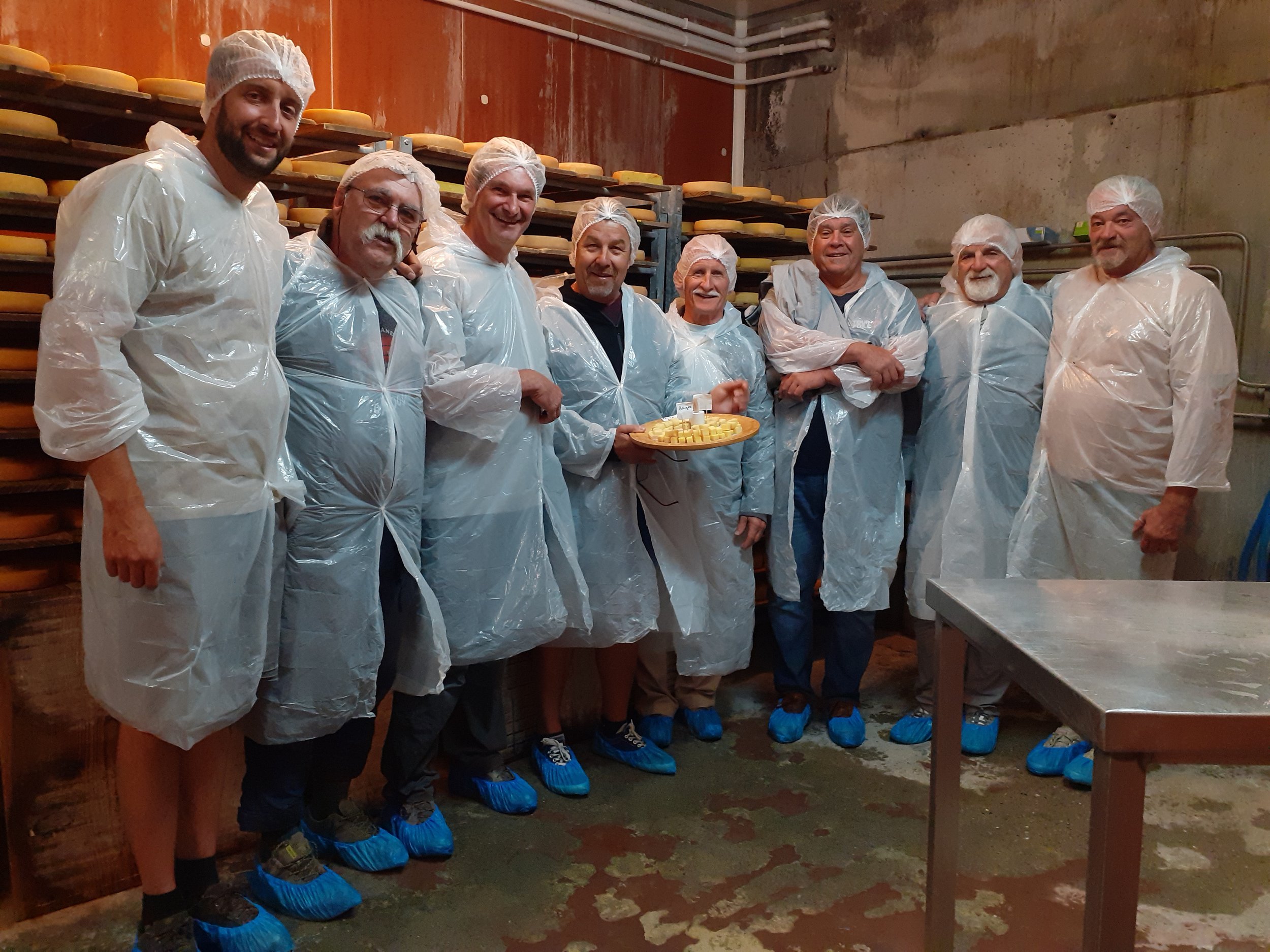 Groupe de personnes en vêtements de protection dans une fromagerie, avec des étagères de fromages derrière eux, certains souriant, un homme tient une assiette de fromages.