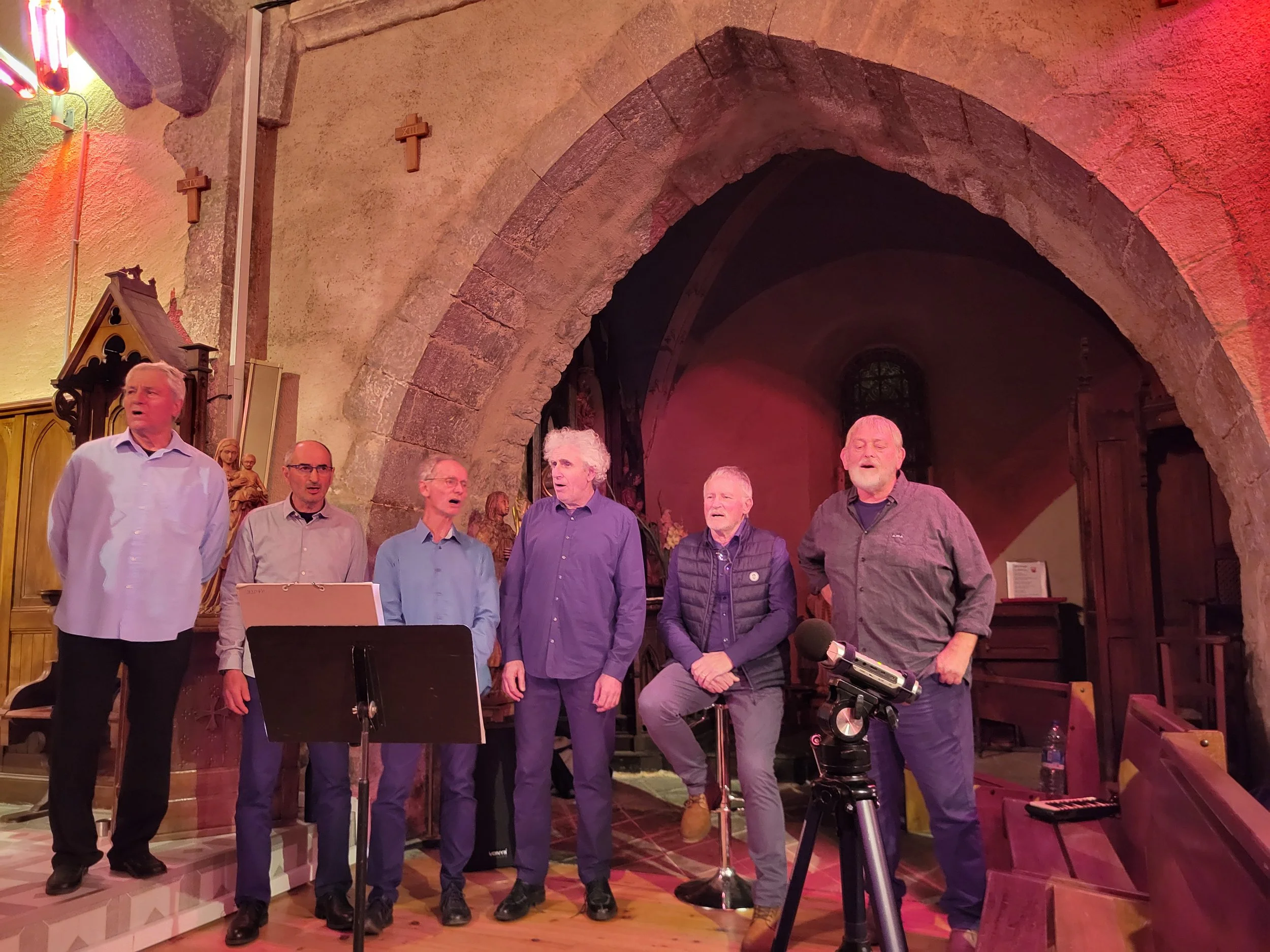 Groupe de sept hommes chantant dans une église, certains avec des partitions, devant un autel avec des statues religieuses.