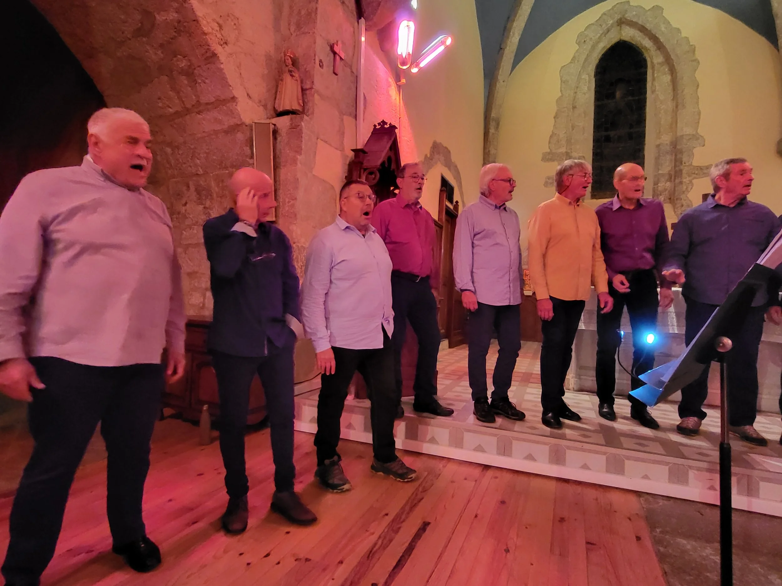 Un groupe d'hommes chantant dans une église, certains avec la bouche ouverte, vêtus de chemises colorées, sous un éclairage chaud, avec des vitraux et des statues en arrière-plan.