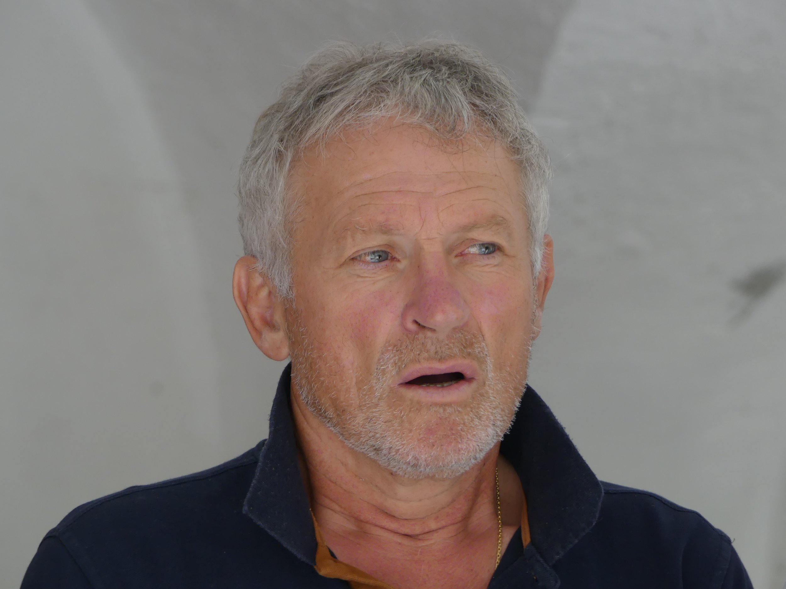 Un homme âgé avec des cheveux gris et une barbe, portant une veste noire et un col relevé, a une expression surprise ou choquée.