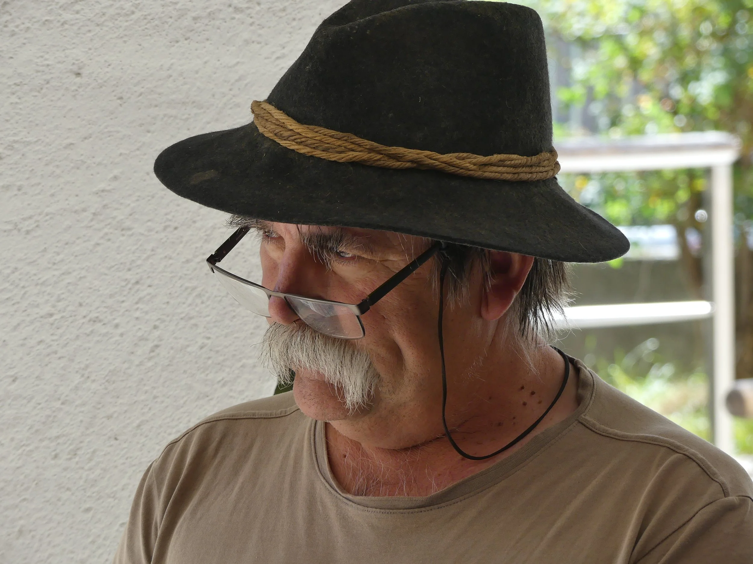 Un homme âgé avec une moustache blanche porte un chapeau noir avec un cordon enroulé autour, des lunettes sur le nez, et un t-shirt beige, regardant vers le bas, en extérieur près d'un mur blanc avec des arbres verts en arrière-plan.