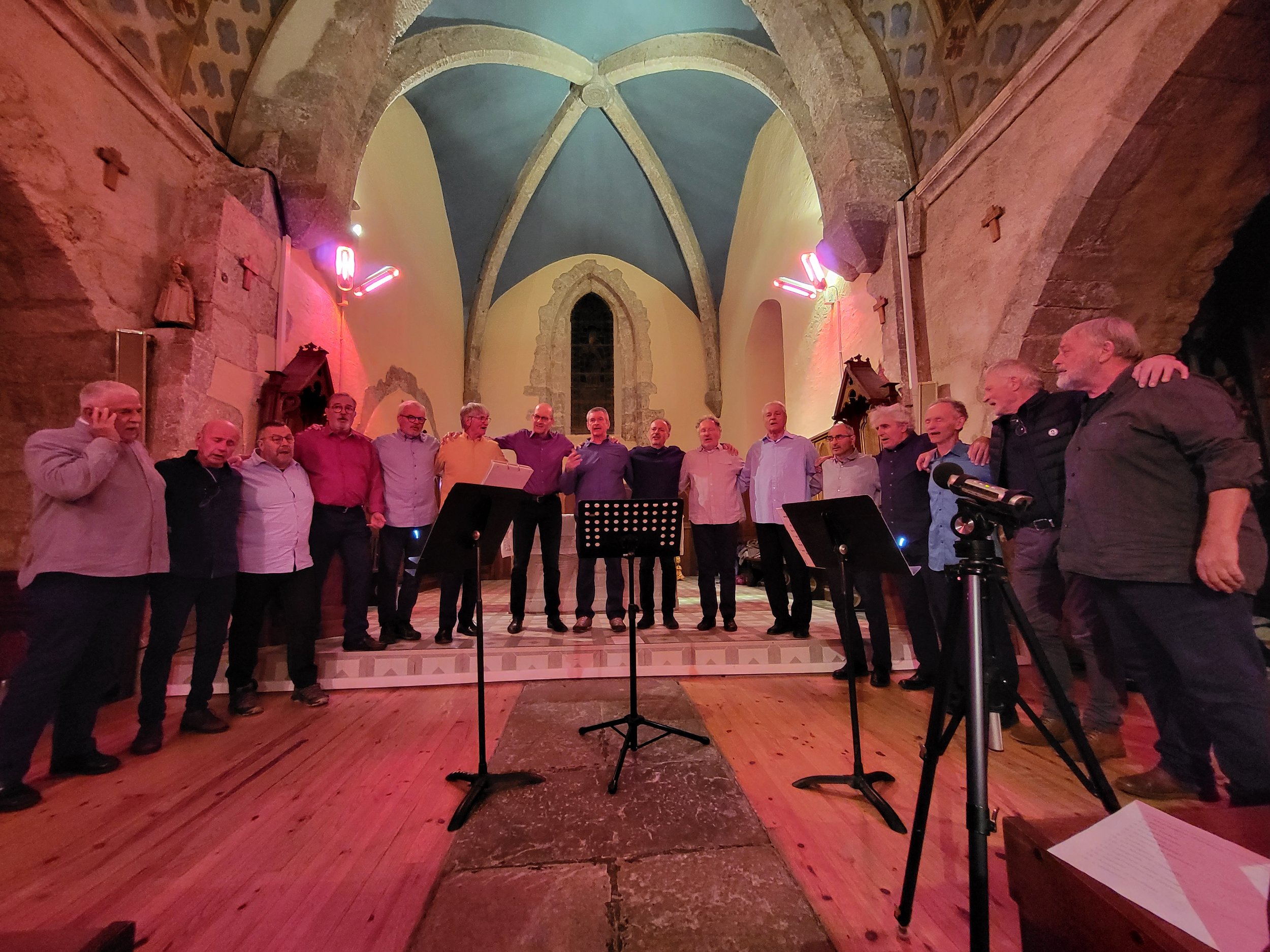 Un groupe de personnes, probablement un chœur, se tient sur une scène dans une ancienne église ou cathédrale. La scène a un plafond voûté avec des éléments artistiques, et des microphones sont placés devant eux. L'éclairage est chaud et suspendu au p