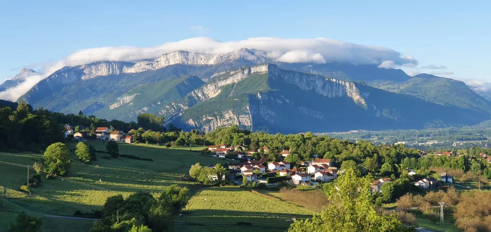 Paysage avec des montagnes, des collines verdoyantes, des maisons et des arbres en premier plan, et des montagnes en arrière-plan sous un ciel partiellement nuageux.
