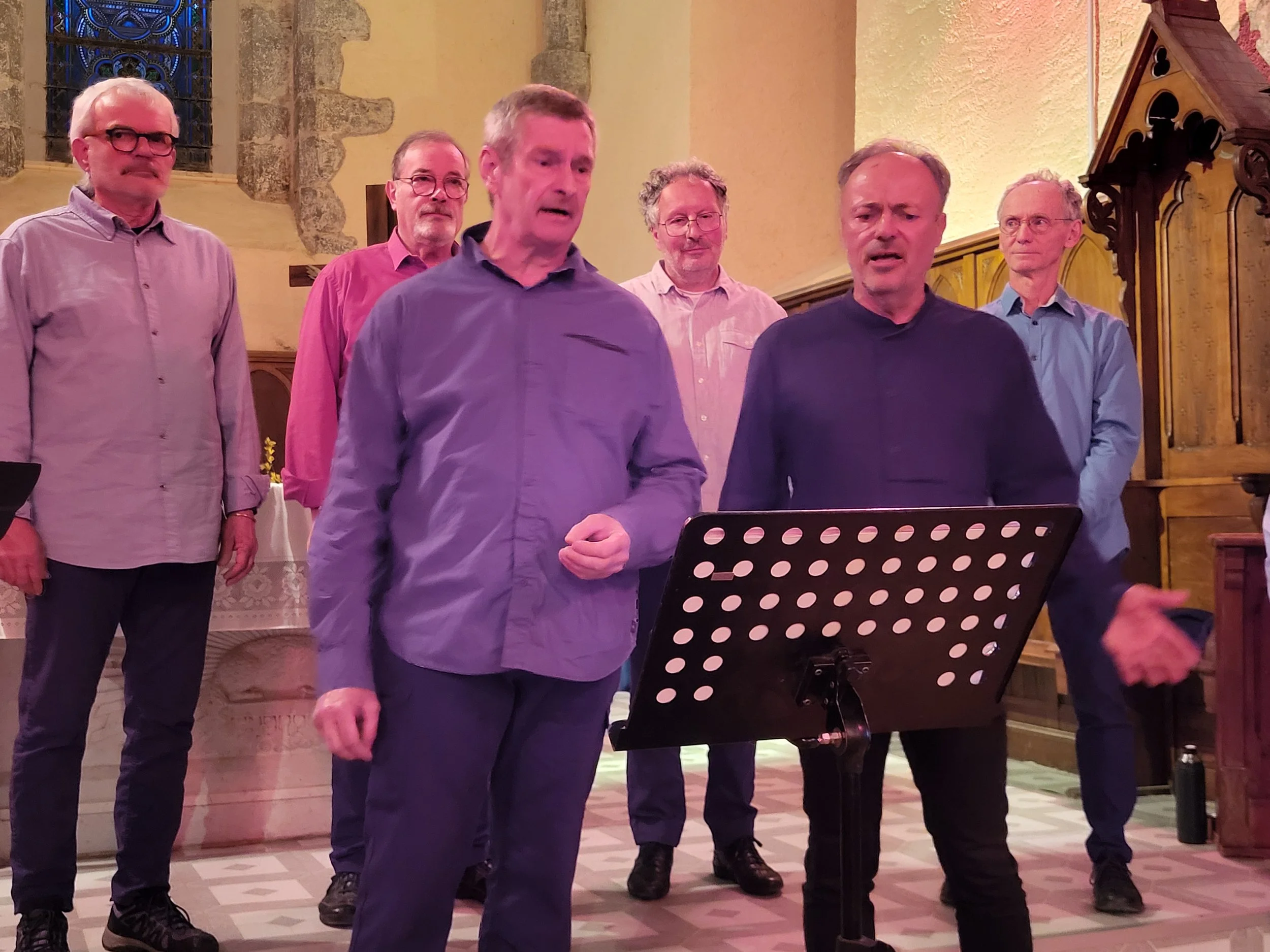 Un groupe de sept hommes chanteurs dans une église. Deux hommes sont au premier plan, un parle et l'autre chante avec une partition. Quatre autres hommes se tiennent derrière eux. La scène est éclairée chaudement, avec un autel visible à l'arrière, d