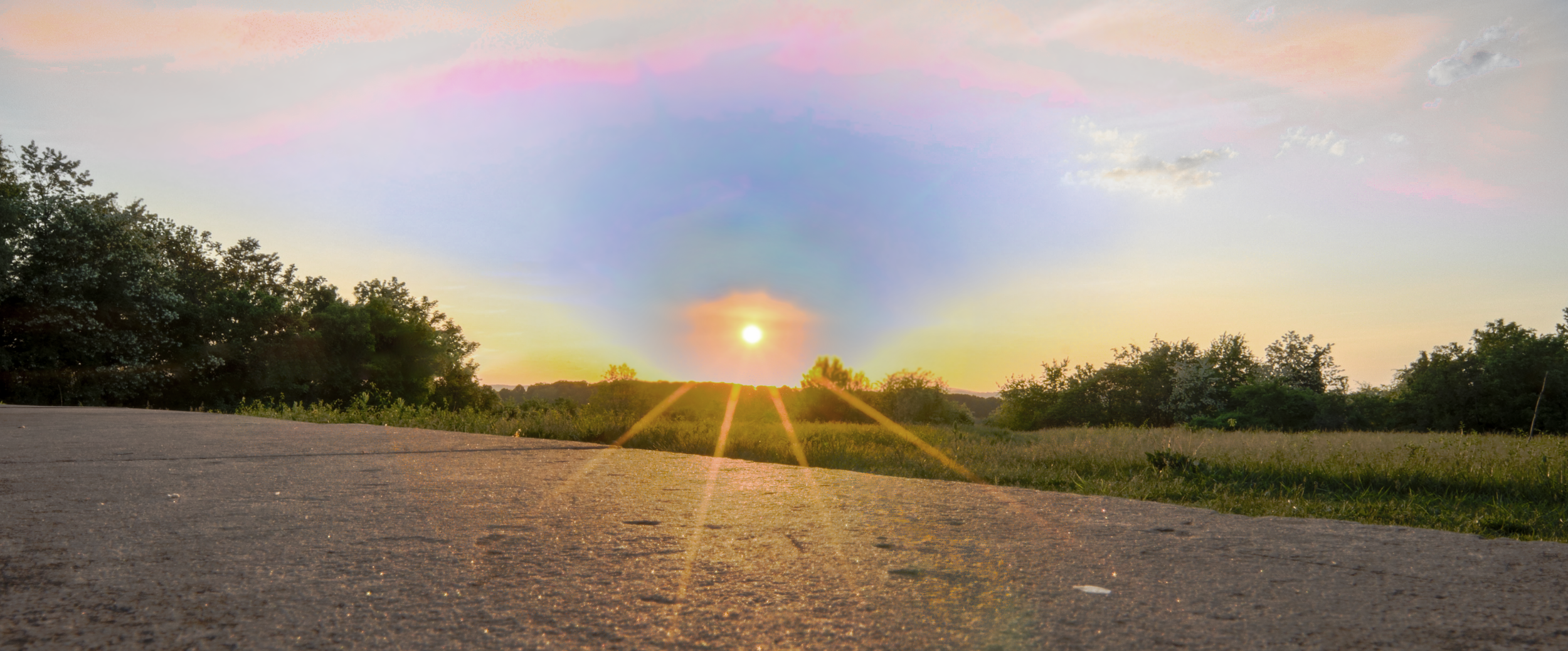 A sunset over a rural landscape with a dirt road in the foreground, green trees and grass, and pink, purple, and orange hues in the sky.