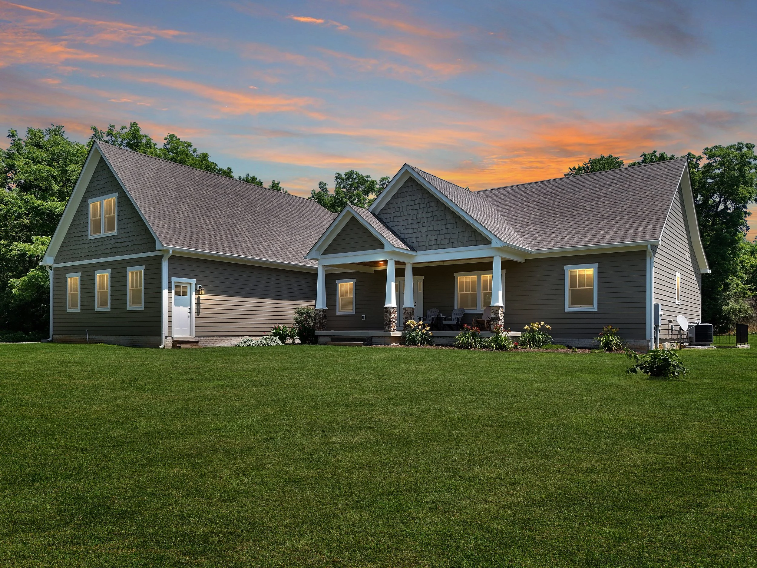 A house with beige siding and a gray roof, illuminated from within, during sunset with a colorful sky, green lawn, and trees in the background.