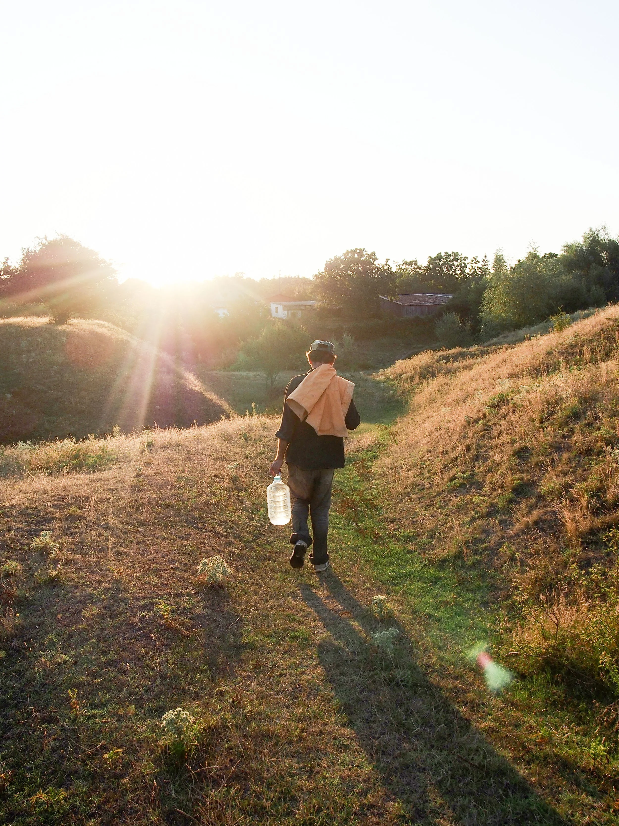 A person walking on a dirt trail in a rural area during sunset, carrying a water bottle, with houses and trees in the background.