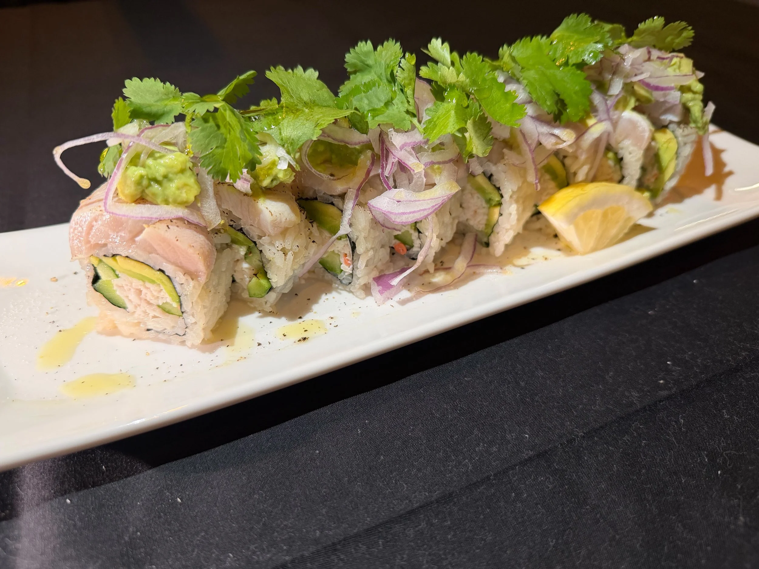 A platter of sushi rolls topped with cilantro, thinly sliced onions, and a piece of lemon, served on a rectangular white plate.
