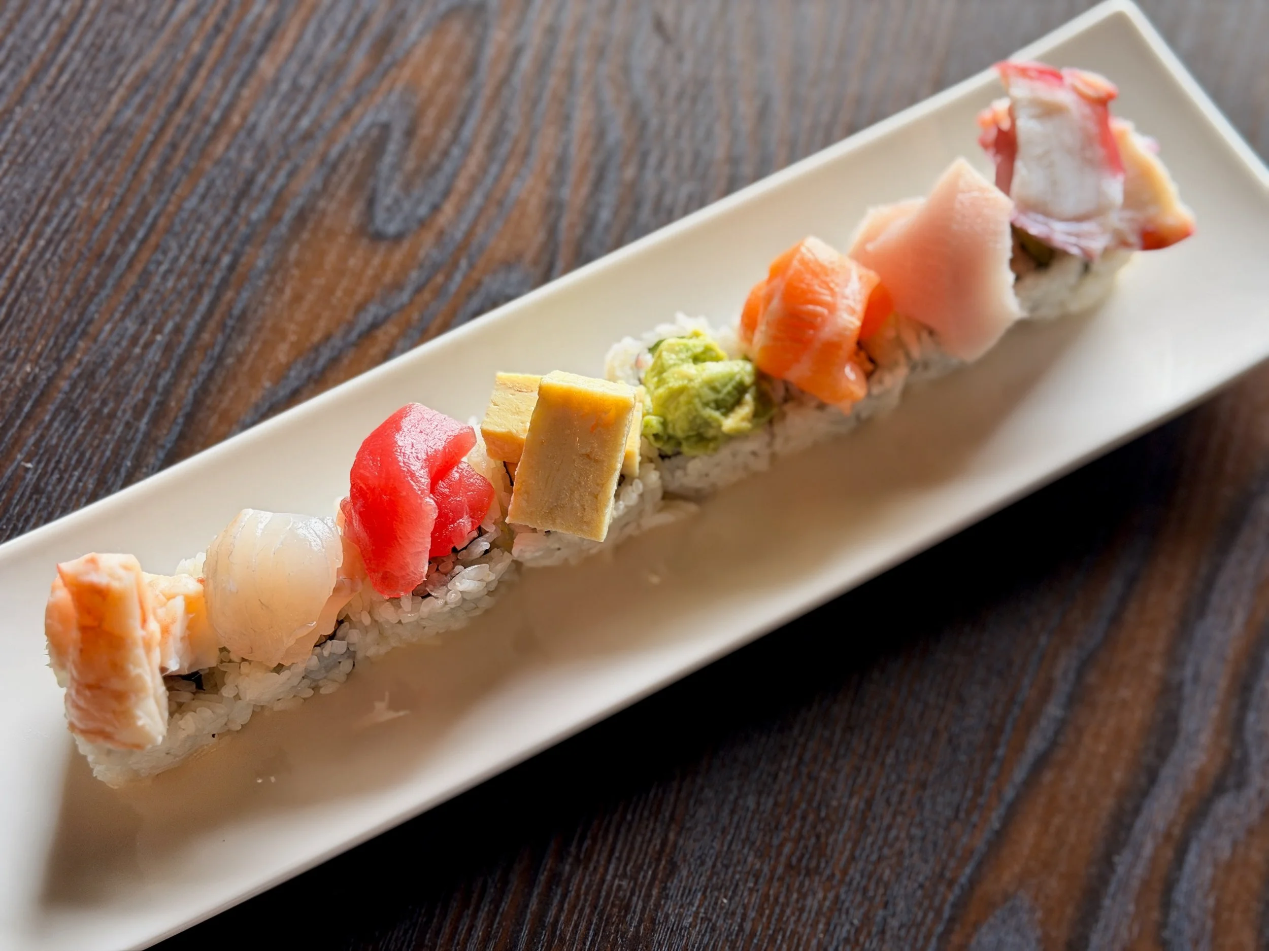 A close-up view of a sushi roll on a rectangular white plate, with various toppings such as fish, avocado, and crab meat, placed on a wooden table.