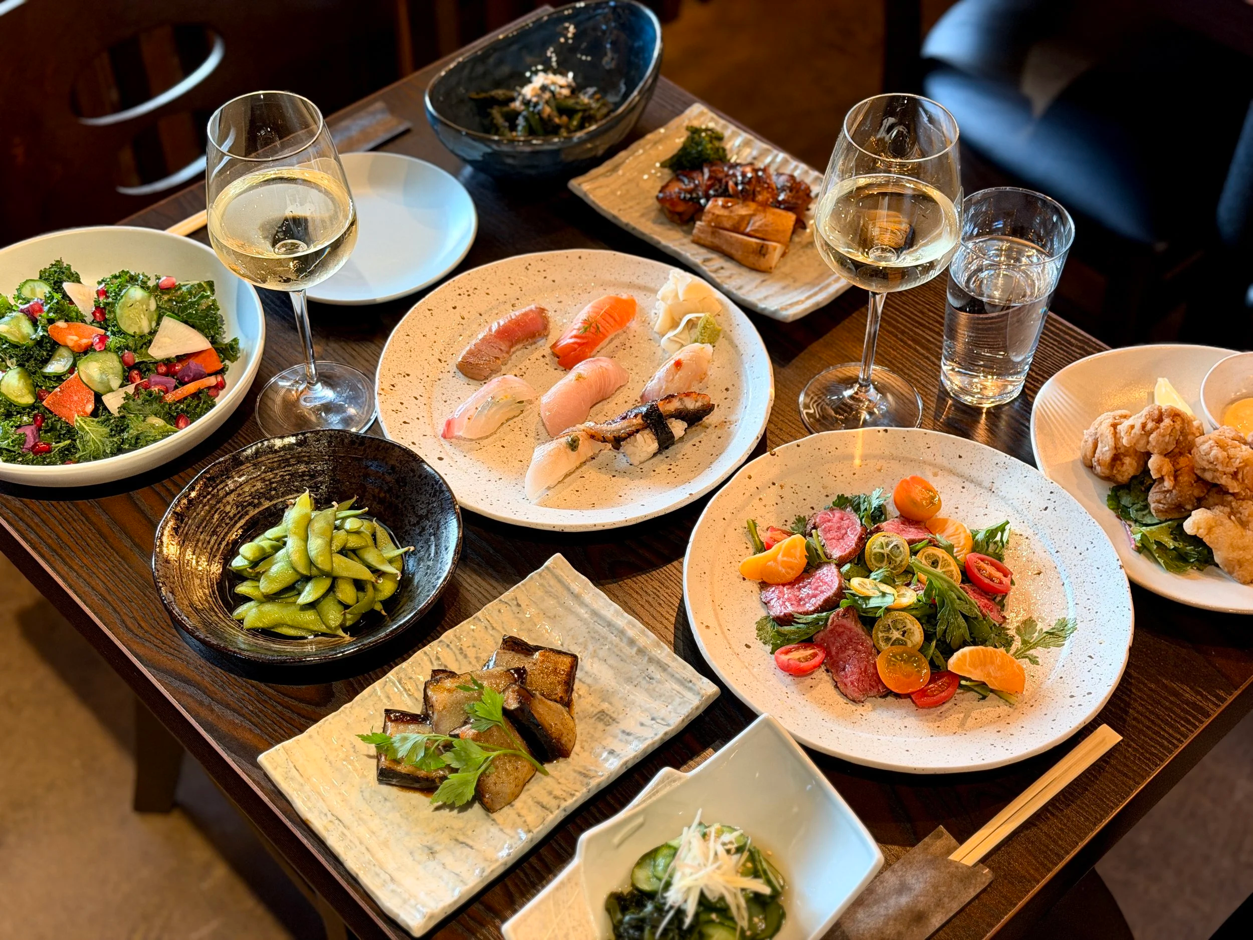 A table with various dishes including salads, sushi, edamame, fried chicken, grilled vegetables, and wine glasses of white wine, set in a restaurant.