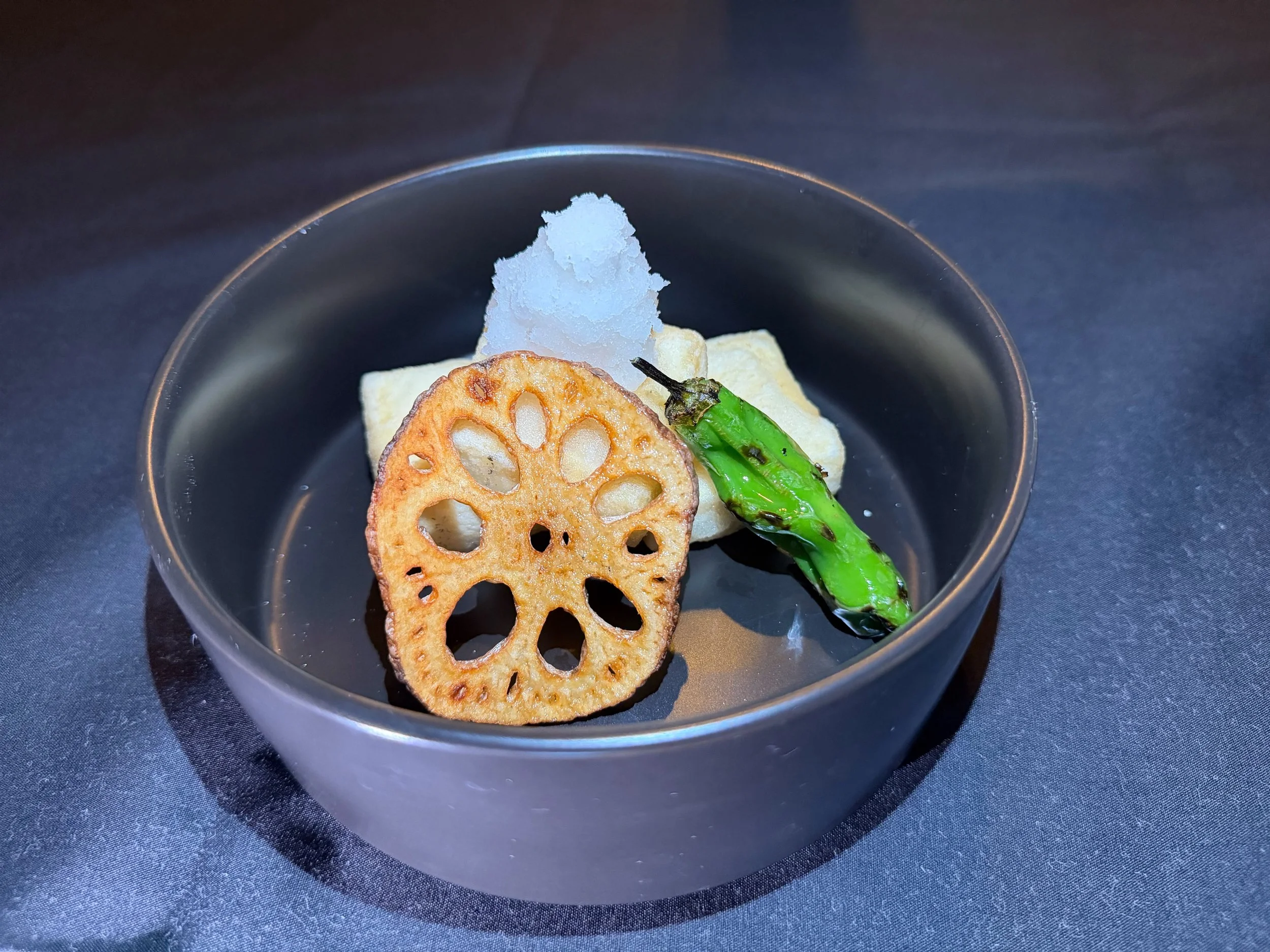 A black bowl contains a piece of fried lotus root, a green chili pepper, a small portion of grated daikon radish, and a piece of tofu, arranged on a dark surface.